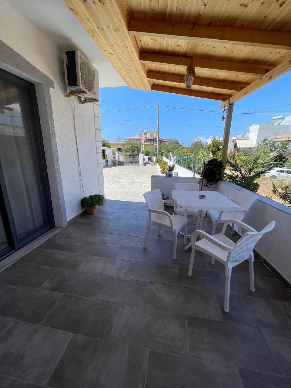 A covered patio area features a modern table surrounded by four white chairs, set against a backdrop of clear blue skies. Potted plants are placed on the tiled floor, enhancing the outdoor space that opens towards a quiet street.