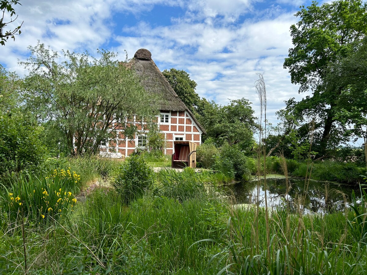 A charming thatched-roof house is surrounded by lush greenery and tall grasses. A serene garden pond reflects the sky, while vibrant yellow flowers add color to the landscape. Trees provide additional shade around the property, enhancing the tranquil setting.