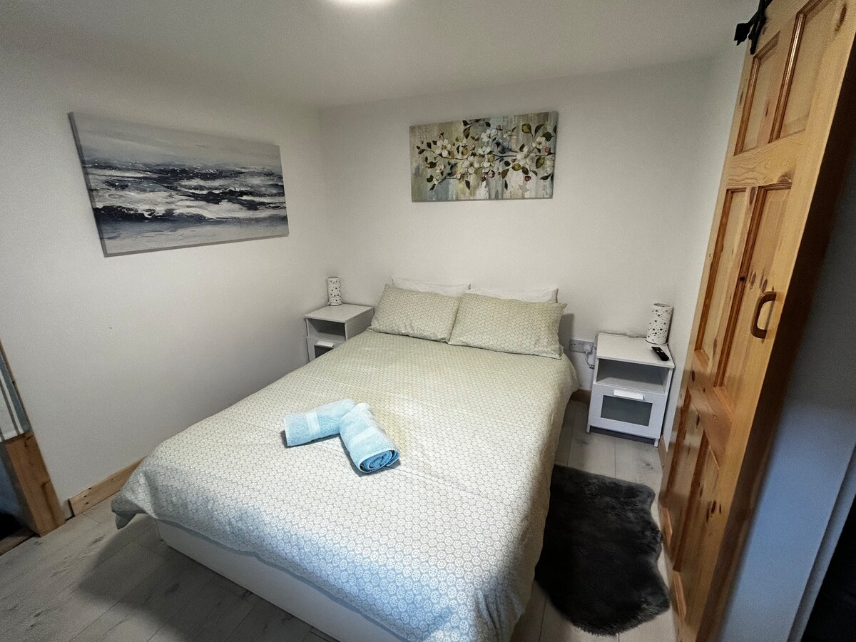 A cozy sleeping area is furnished with a comfortable double bed, covered with a patterned duvet and complemented by two bedside tables. Soft blue towels are neatly arranged on top of the bed. A floral artwork adorns the wall above, enhancing the tranquil atmosphere.