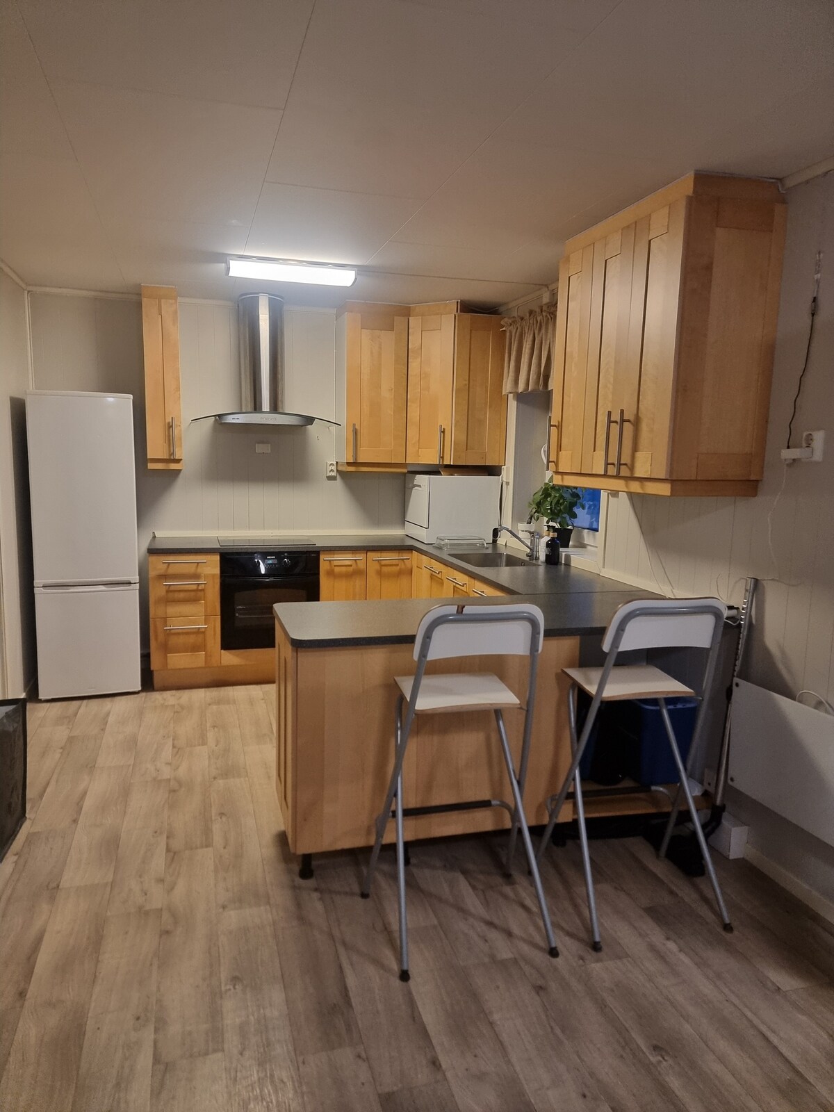 The kitchen features wooden cabinetry and a modern layout. A refrigerator and oven are integrated into the design. Two bar stools are positioned at a small table, while natural light enters through the window, complementing the neutral tones of the space.