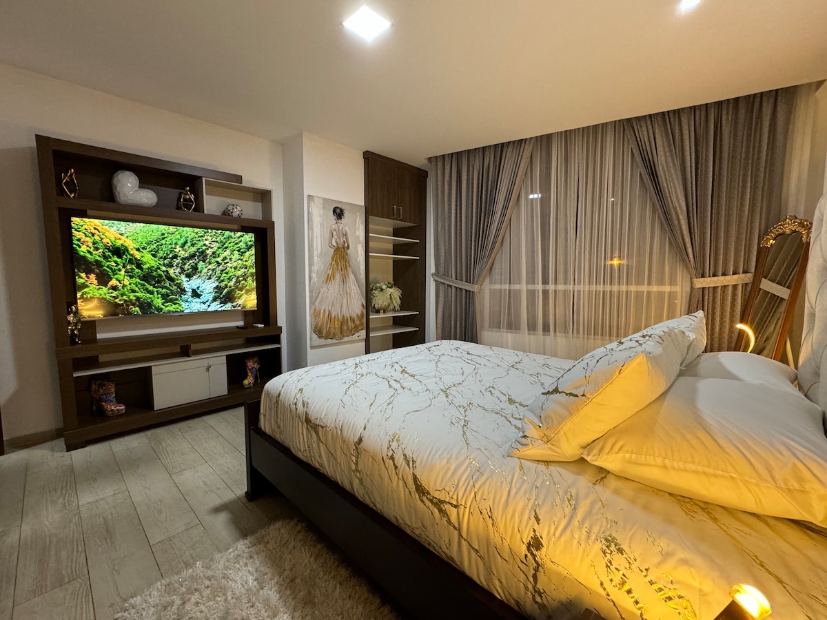 The spacious bedroom features a large bed with adorned pillows and a decorative headboard. A modern entertainment center houses a flat-screen TV, framed by light curtains that filter soft natural light. The room is furnished with minimal shelving, contributing to a clean, open feel.