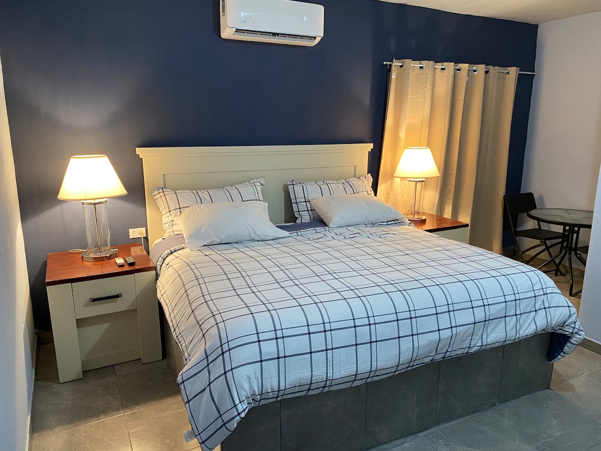 A spacious sleeping area is furnished with a large bed dressed in a plaid duvet. Two bedside tables hold lamps providing soft lighting. A small circular table and chairs are visible in the corner, with an air conditioning unit mounted above, enhancing comfort throughout.