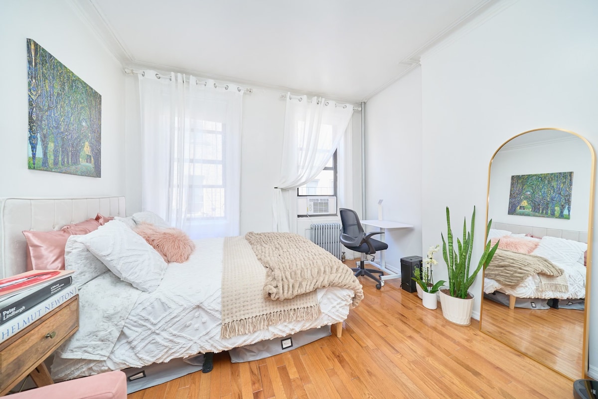 A bright, airy studio features a spacious bed with soft white bedding and decorative pillows. Natural light filters through large windows, illuminating the room. A desk and chair are positioned near a window, while plants and artwork add a touch of nature and color to the space.