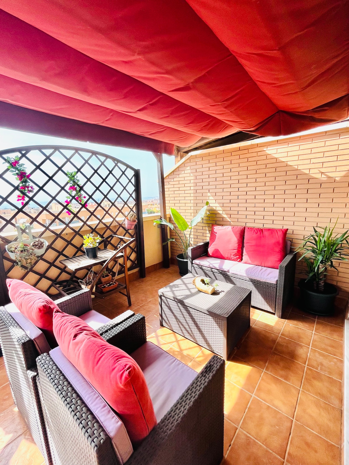 A terrace area is presented with several seating options, including two armchairs and a small coffee table. Cushions in bright red add a pop of color, while potted plants and decorative wall elements create a welcoming environment. Sunlight filters through, enhancing the space's brightness.