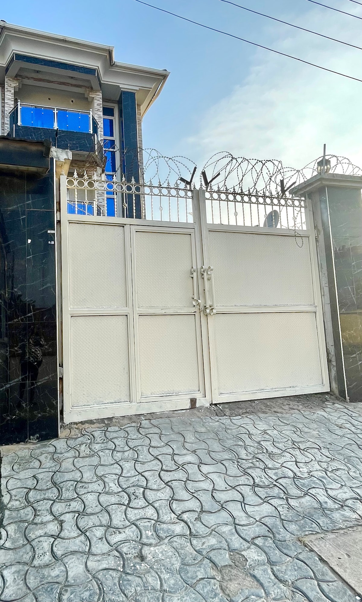 A white metal gate is secured with a padlock, featuring a design of intertwined bars and barbed wire along the top. The gate is framed by dark stone walls, with a patterned cobblestone pathway leading towards it. The sky above is clear, reflecting daylight.