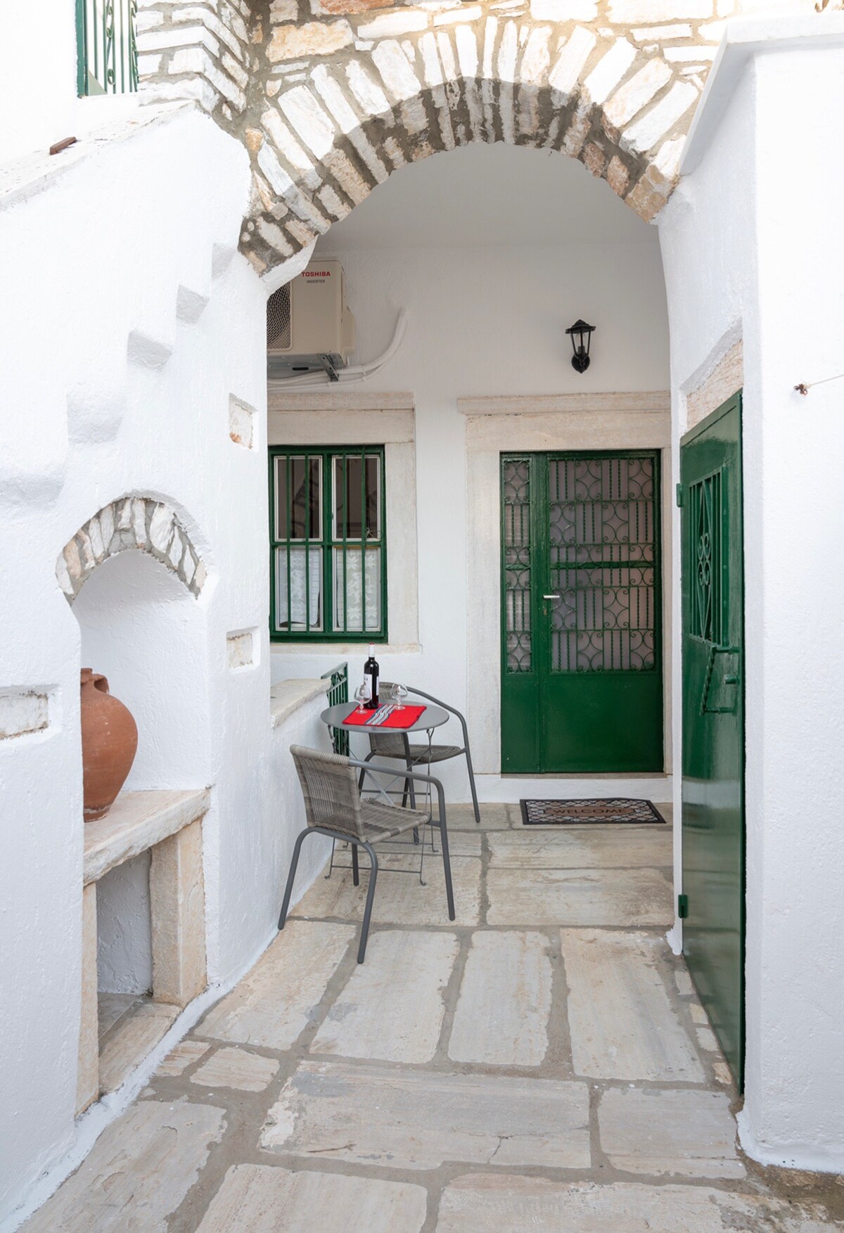 A charming courtyard is framed by whitewashed walls and a stone archway, showcasing a small table set for two with a bottle of wine. Green doors and windows provide a pop of color against the neutral stone floor, enhancing the welcoming ambiance.