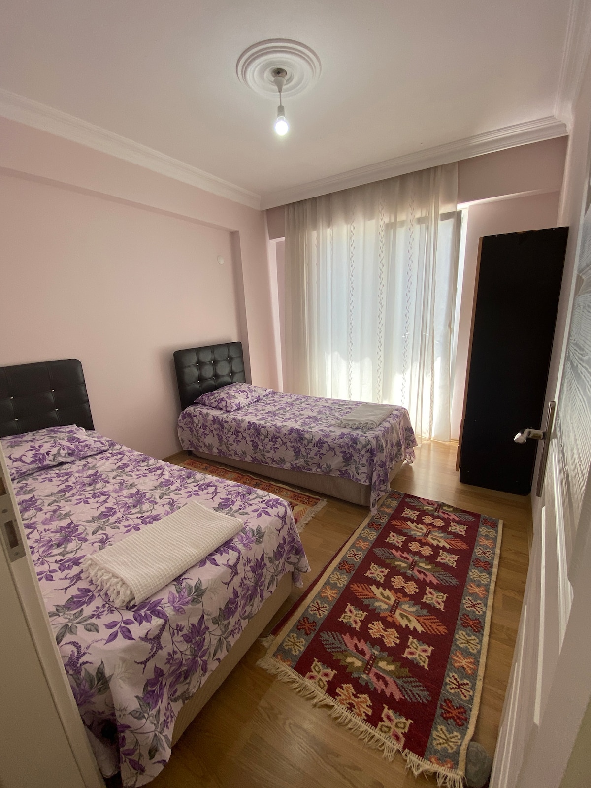 A room features two beds with floral-patterned linens and towels placed neatly on one. Natural light fills the space through a large window, while a dark wardrobe stands against the wall. A colorful rug adds warmth to the wooden flooring.