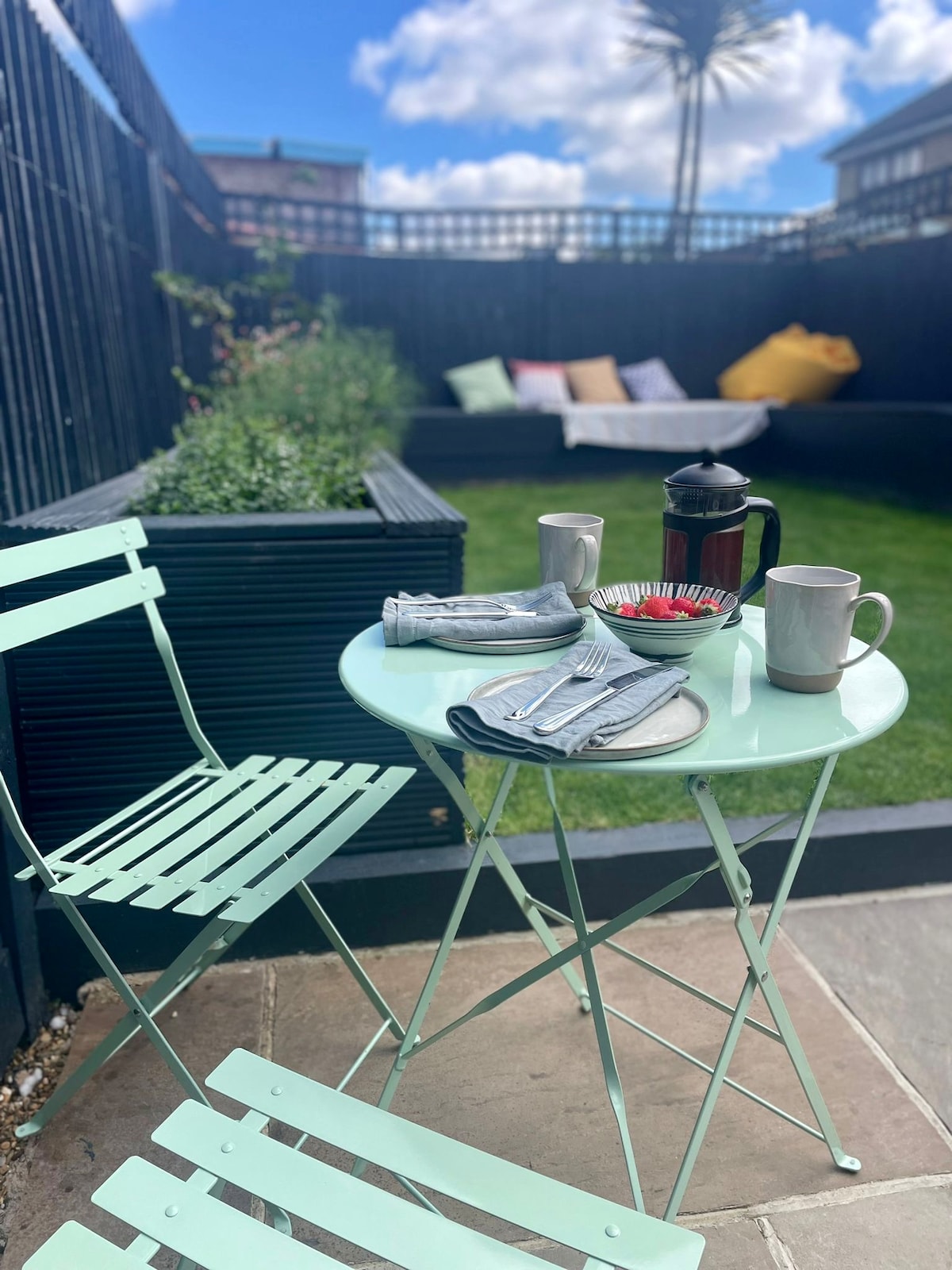 A small outdoor dining setting is presented in the garden, featuring a mint green table paired with matching folding chairs. Two mugs and a coffee press are positioned on the table, accompanied by a plate and utensils. The background includes a seating area bordered by greenery.