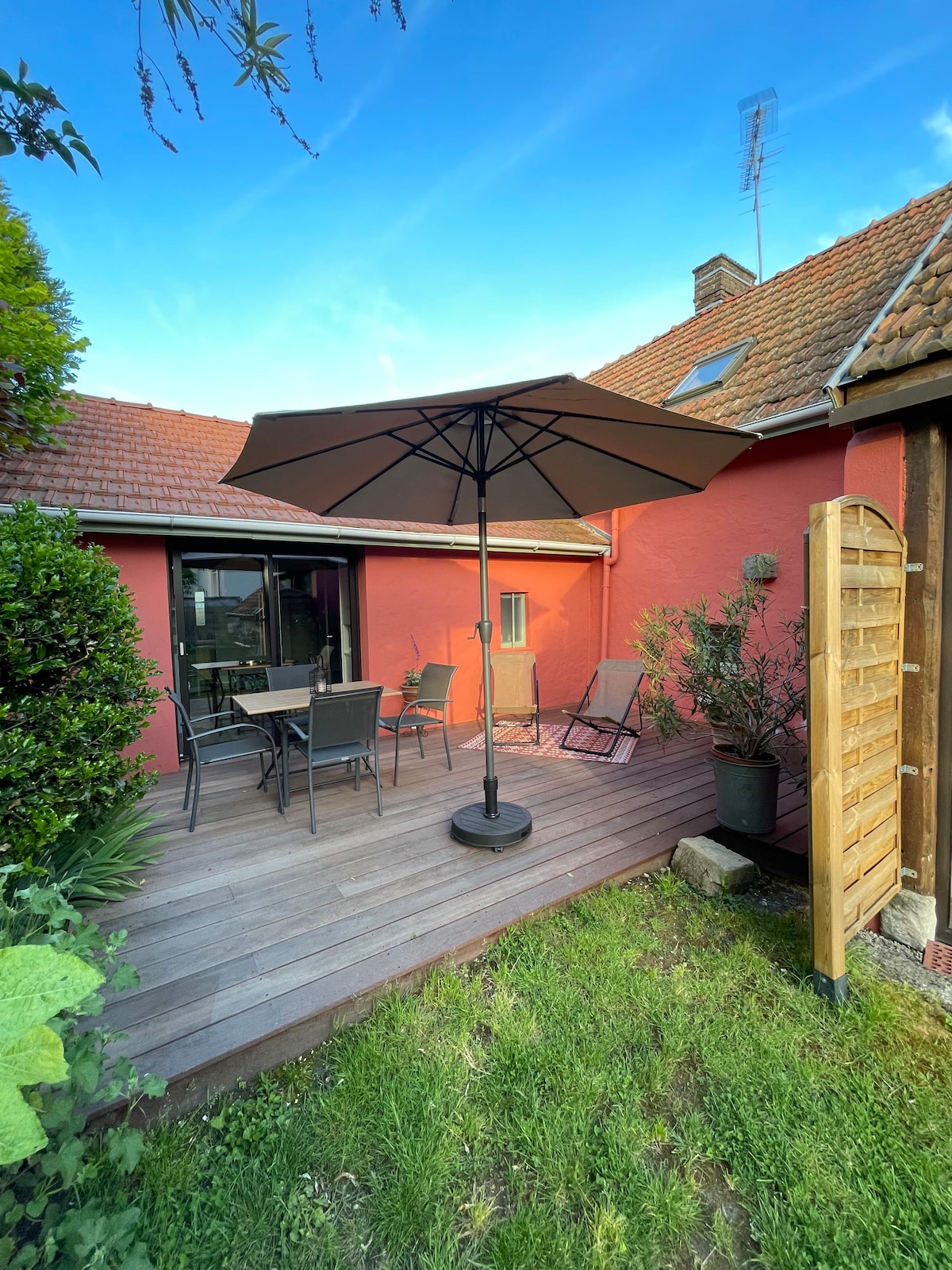 A private terrace is showcased with wooden decking and seating arrangements that include a table and chairs under a large umbrella. Lush greenery and potted plants frame the area, providing a serene space for relaxation.