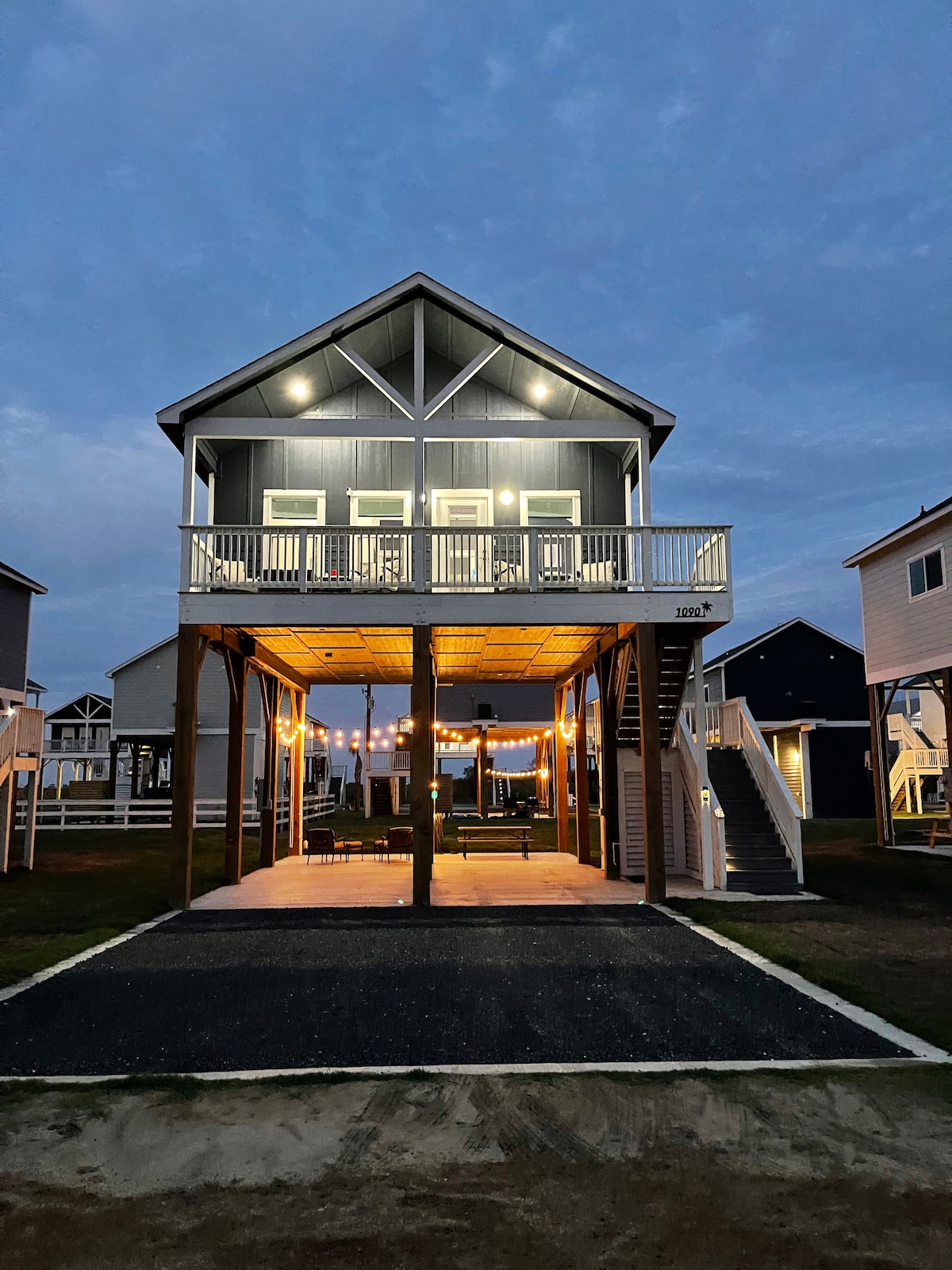 The villa is shown in the evening, illuminated by outdoor lights that enhance its welcoming appearance. Spacious decks are visible on both levels, with a clean area beneath supported by pilings. Nearby villas can be seen in the background, showcasing the coastal neighborhood.