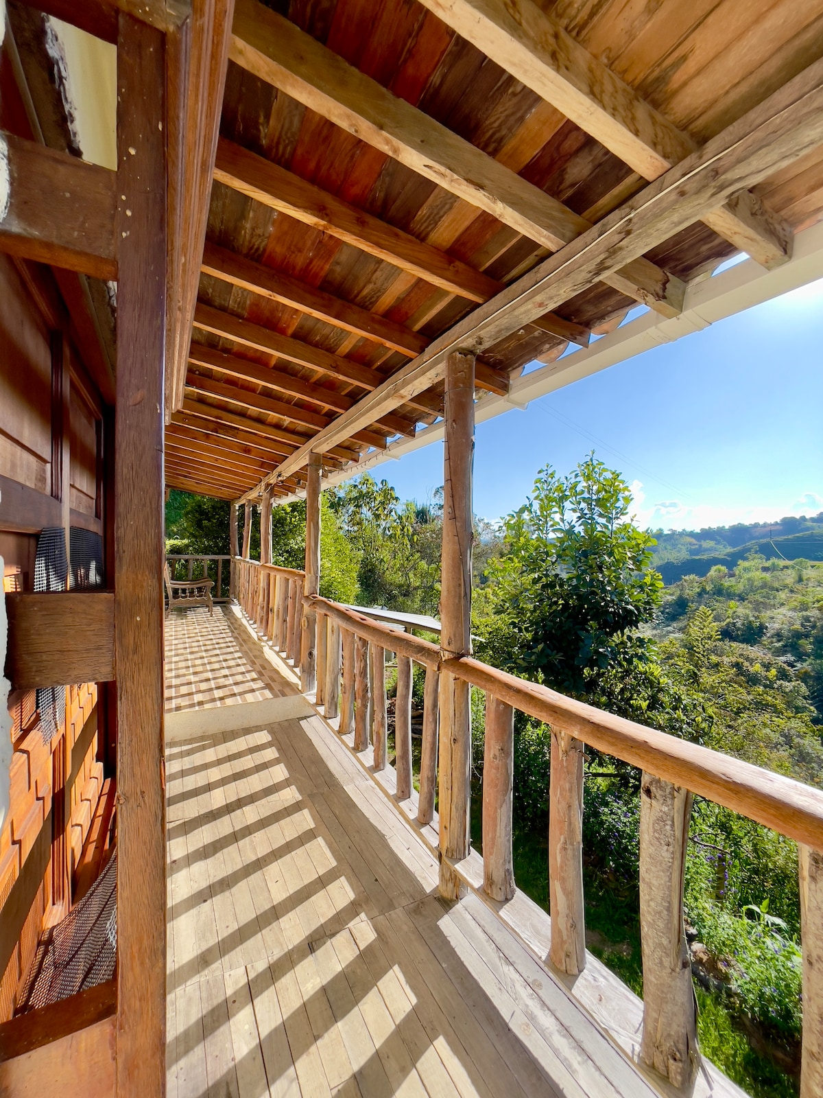A wooden balcony extends from the cabin, featuring rustic railings and a wooden floor. Natural light floods the space, illuminating the surrounding greenery and distant hills beyond. The open view invites relaxation and connection with nature.