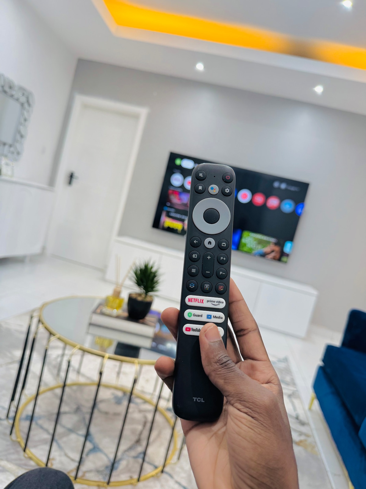 A hand holds a remote control in the foreground, with a modern living room visible in the background. A large television displays a screen with various application icons. Bright lighting and a decorative coffee table complement the contemporary decor.