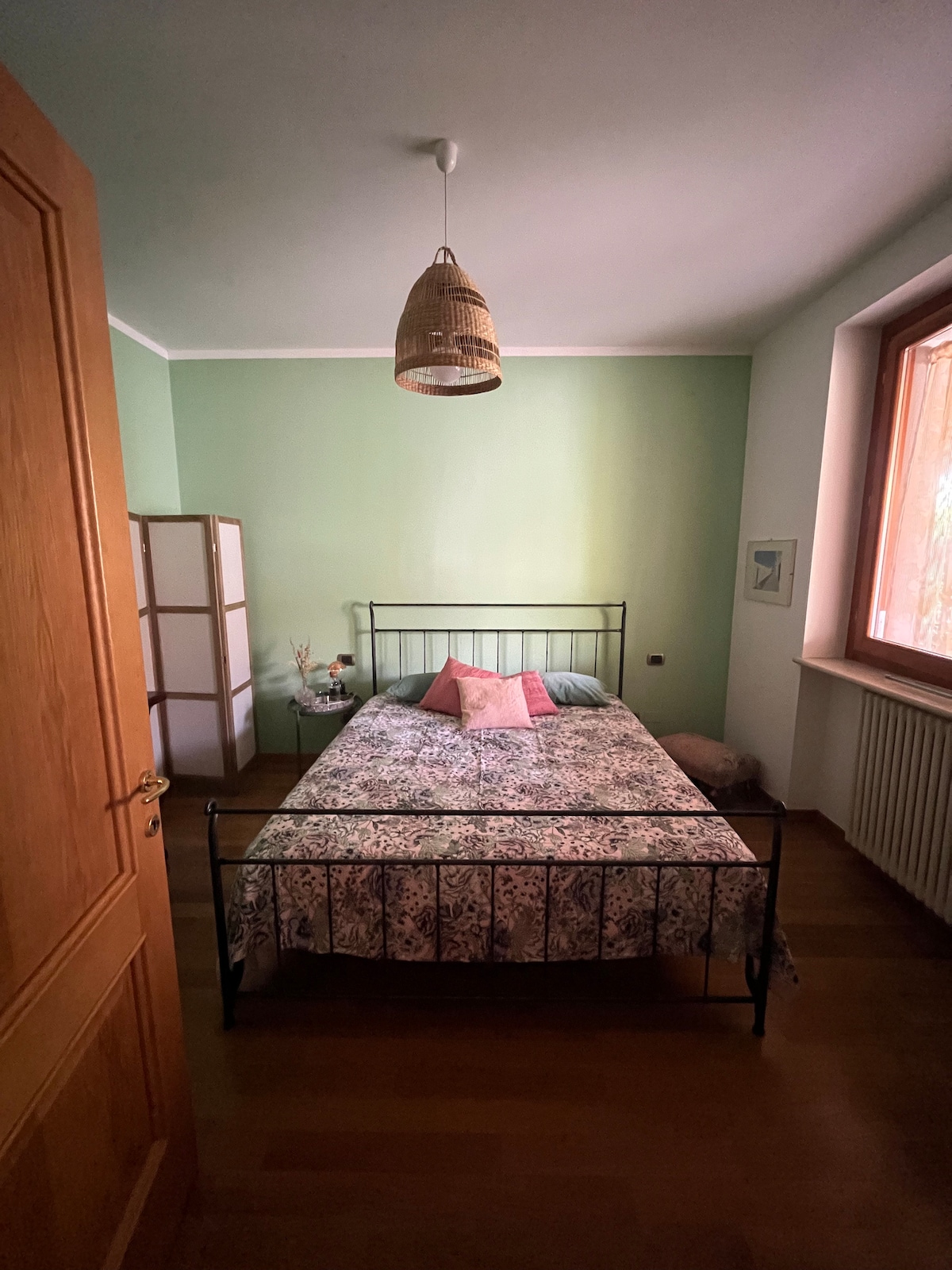 A cozy bedroom is presented, featuring a spacious bed with a patterned bedspread and two decorative pillows. A woven light fixture hangs from the ceiling. Natural light streams through a window, illuminating soft green walls. A wooden door leads into the room, enhancing a sense of privacy.