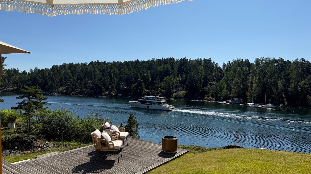A wooden deck overlooks a serene waterway, framed by lush greenery and trees. Comfortable seating is arranged on the deck, providing a view of passing boats. A fire pit sits nearby, enhancing the outdoor experience. The sky is clear and blue.