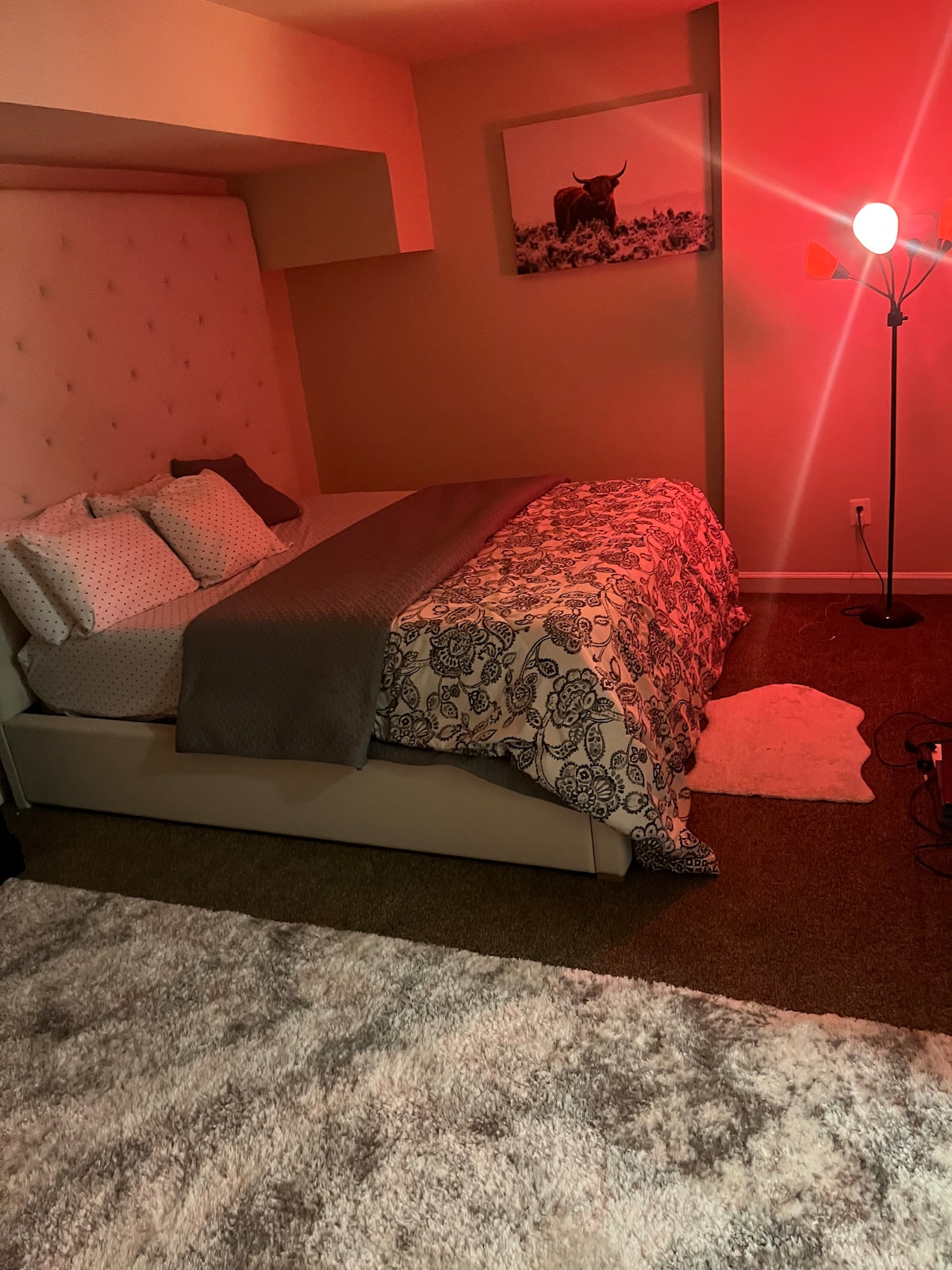 A cozy bedroom features a plush, light-colored rug covering the floor. A modern bed with a tufted headboard is dressed with decorative pillows and a patterned blanket. A floor lamp adds soft light, creating a warm ambiance, while a nature-themed artwork is displayed on the wall.