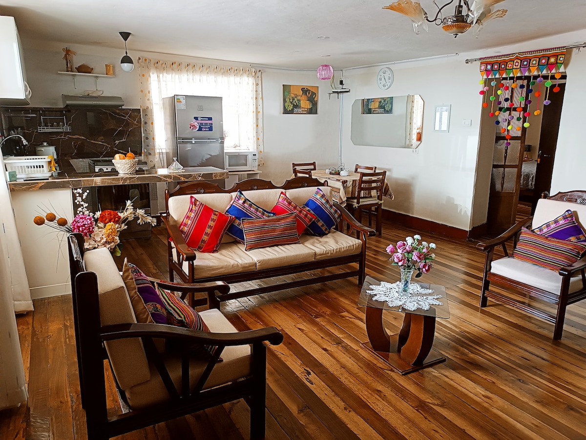 A well-furnished living area is observed, featuring a comfortable seating arrangement with decorative cushions. A glass table occupies the center, while a fully equipped kitchen is visible in the background. Wooden floors reflect light, adding warmth to the space.