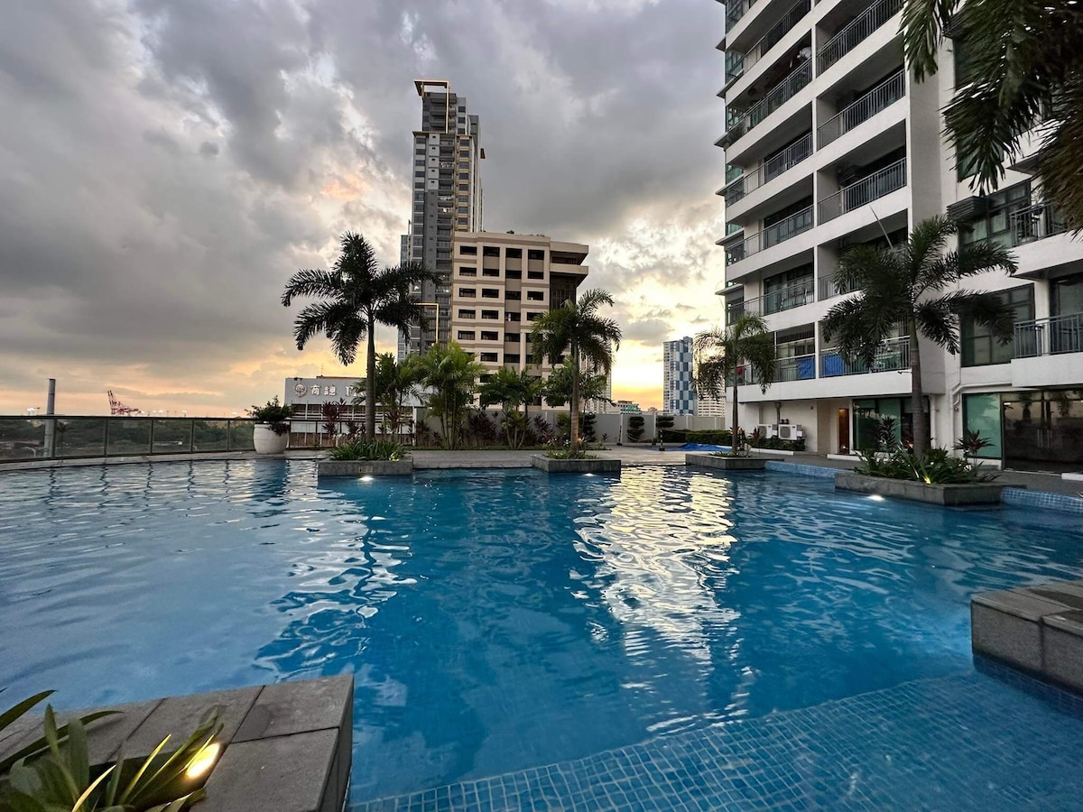 An infinity pool is surrounded by palm trees and modern buildings, reflecting the cloudy sky. Sunlight glimmers on the water's surface, creating a serene atmosphere. Lounge areas with tropical plants are visible along the pool's edge, inviting relaxation.