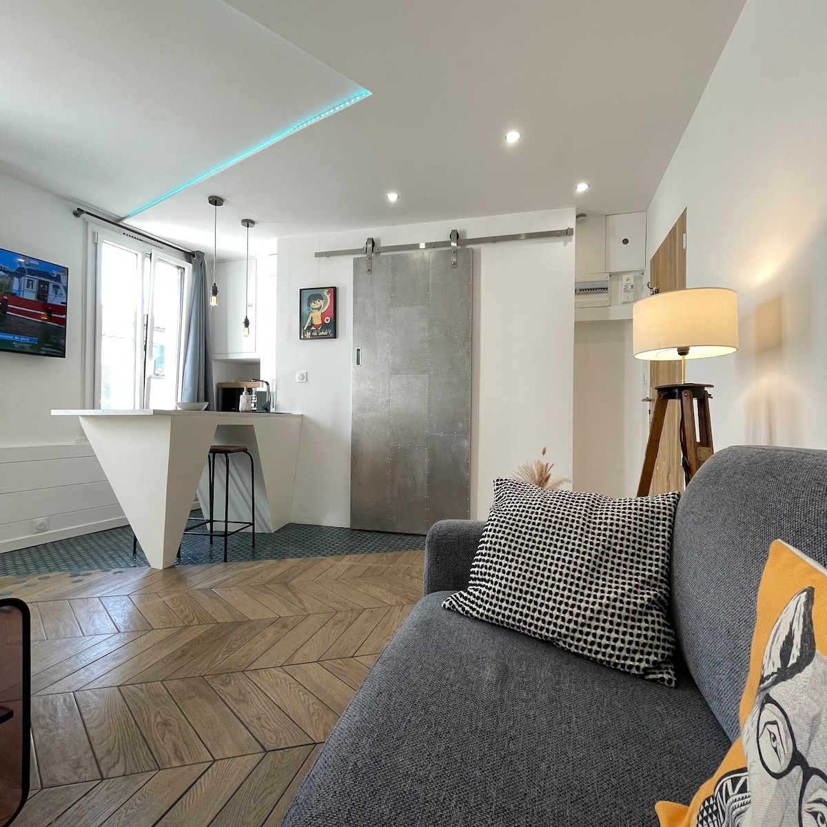 A modern living area is presented, featuring a comfortable gray sofa adorned with patterned cushions. A sleek kitchenette with a bar-style counter is visible in the background, complemented by a decorative wall art piece. Natural light enters through a large window with neutral-colored curtains.