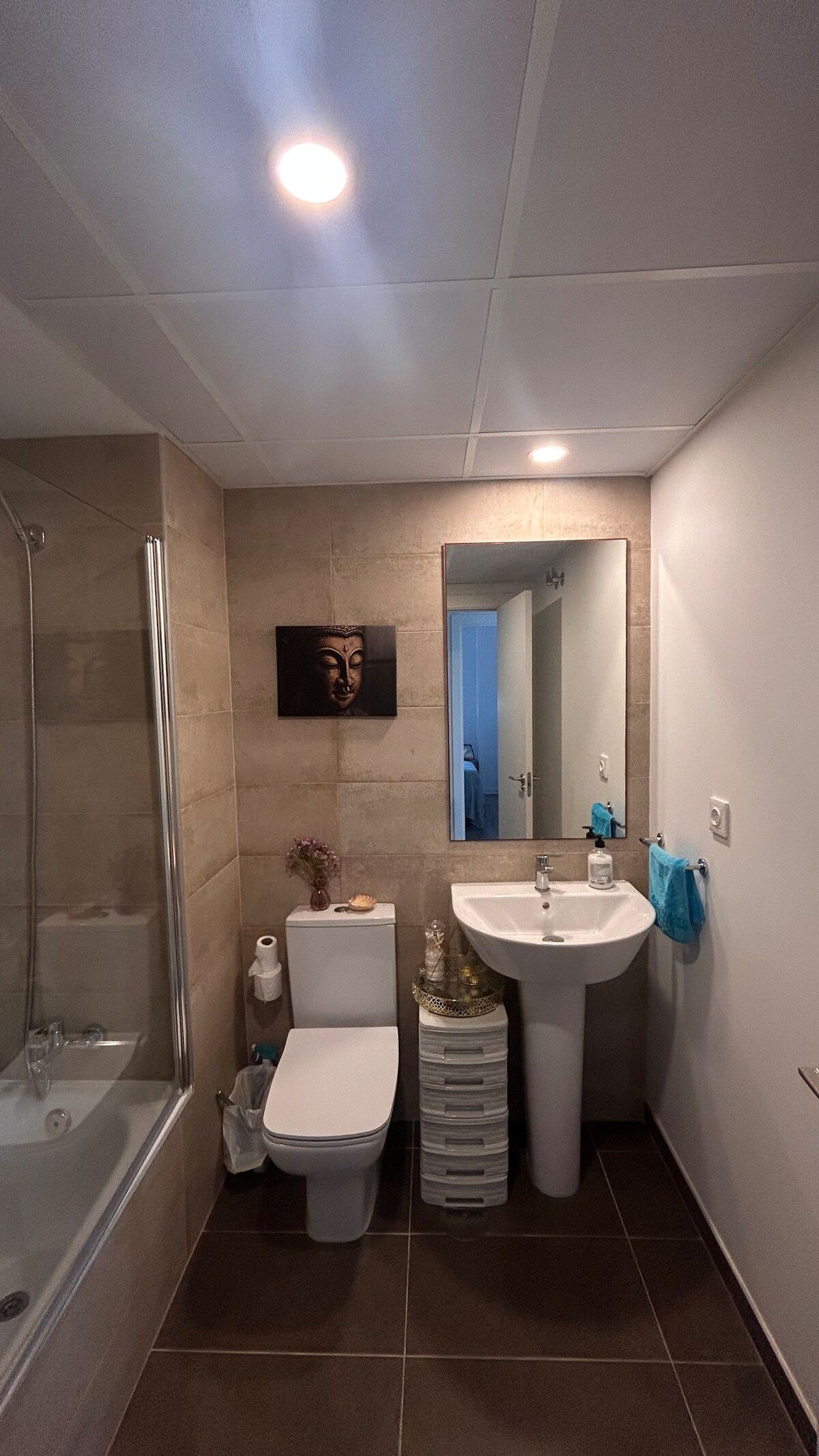 A modern bathroom features a relaxing bathtub alongside a sleek sink and toilet. Soft lighting creates a bright atmosphere. The mirror above the sink reflects the room, while a small television is mounted on the wall, providing convenience. Storage space is offered by a white organizer on the floor.