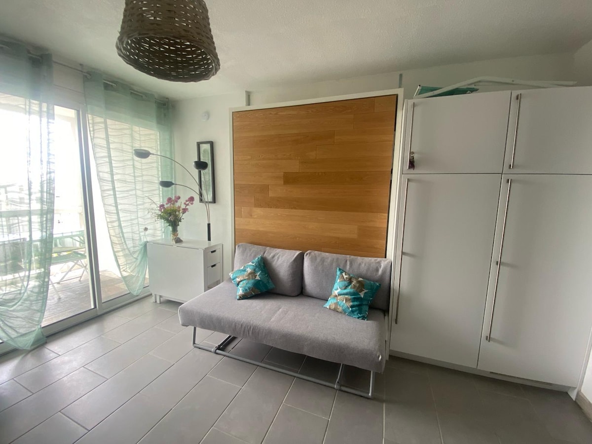 A bright living area features a gray convertible sofa adorned with two decorative cushions. A wooden wall panel serves as a backdrop, complemented by a sleek white cabinet. Large glass doors provide access to a terrace, with light sheer curtains gently swaying in the breeze.