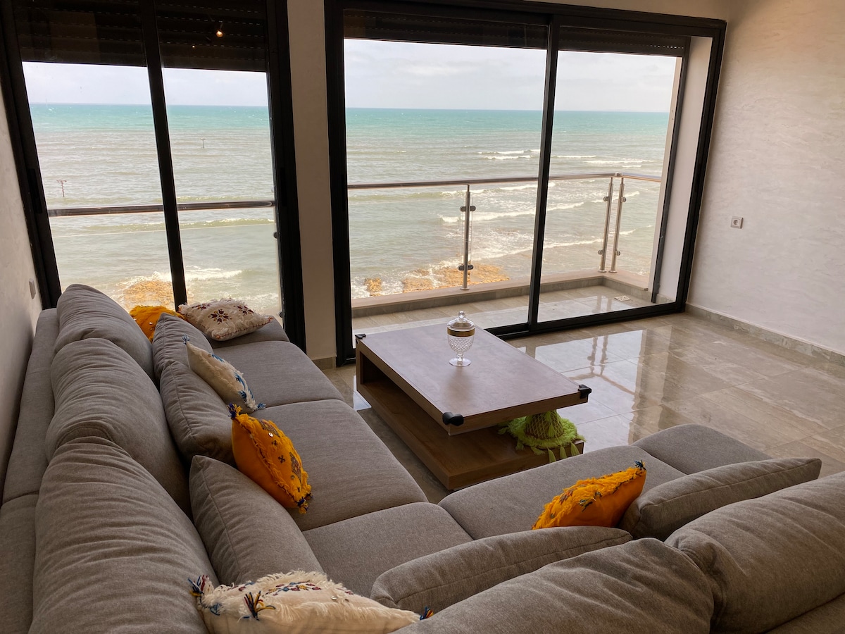 A spacious living area features large glass doors that open to a stunning ocean view. A comfortable gray sectional sofa is adorned with vibrant yellow cushions. A wooden coffee table sits centrally, complementing the serene atmosphere created by the nearby sea.