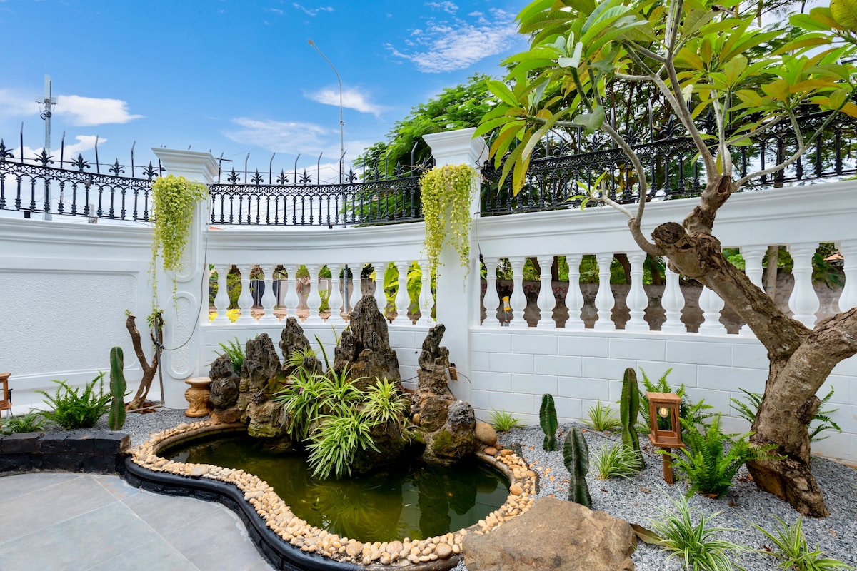 A tranquil outdoor space is showcased, featuring a pond surrounded by carefully arranged stones and lush greenery. Decorative plants cascade from the balustrade above, enhancing the serene atmosphere. The space is complemented by artistic rock formations, creating a harmonious blend of nature and design.