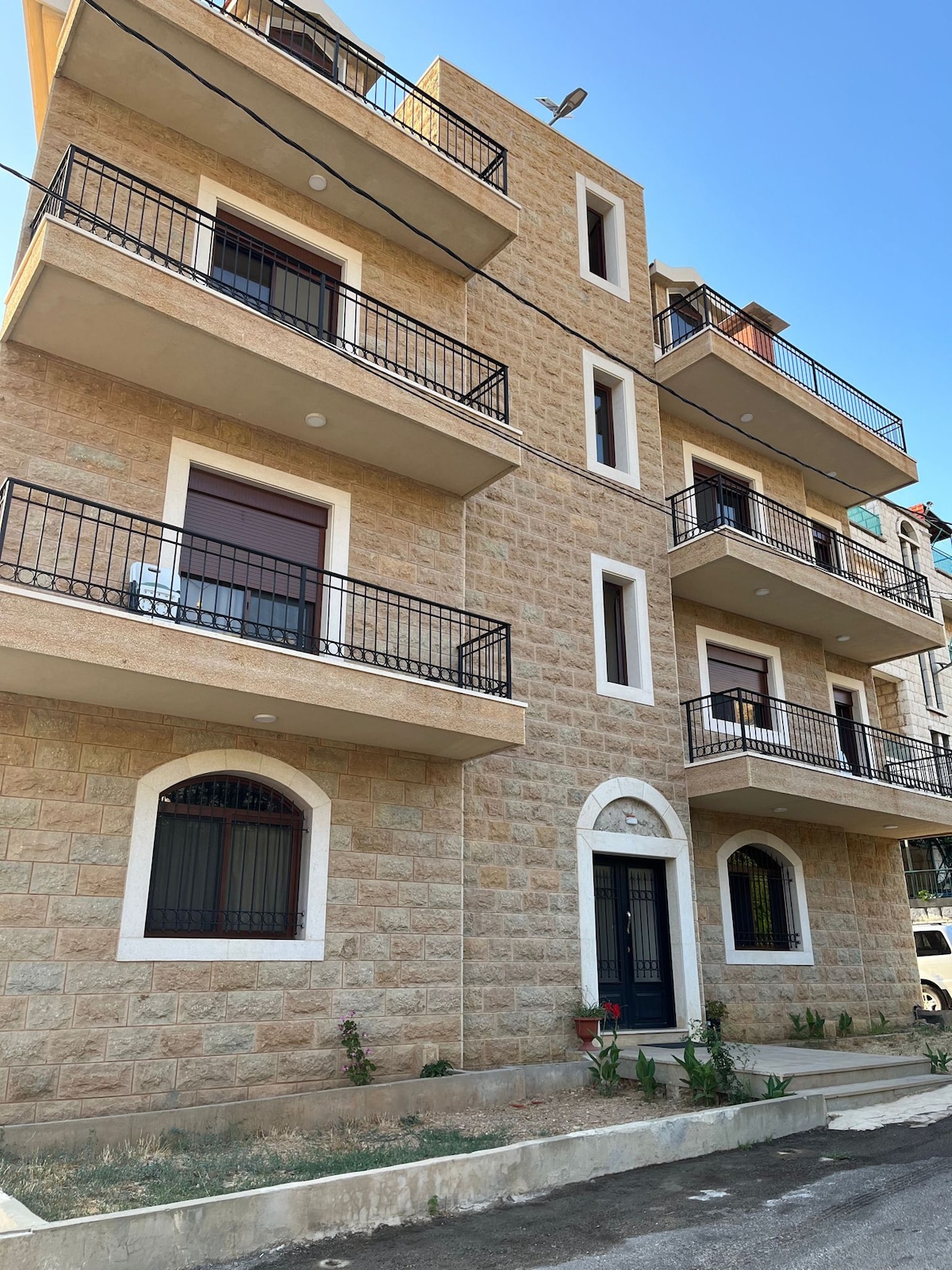 The exterior of a newly finished stone building is showcased, featuring three levels with multiple balconies. Each balcony is surrounded by black railings, and the entrance is marked by a decorative arch. Clear blue skies are visible above, enhancing the inviting appearance.