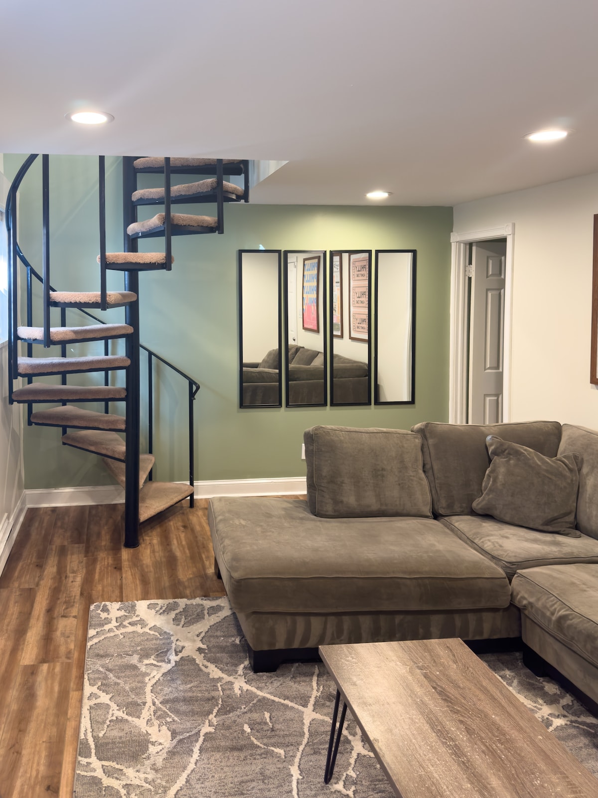 A spacious living area is shown with a large sectional sofa and a wooden coffee table. A spiral staircase leads to the upper floor. Three mirrors are mounted on the wall, reflecting the cozy ambiance of the room, adorned with a neutral color palette.