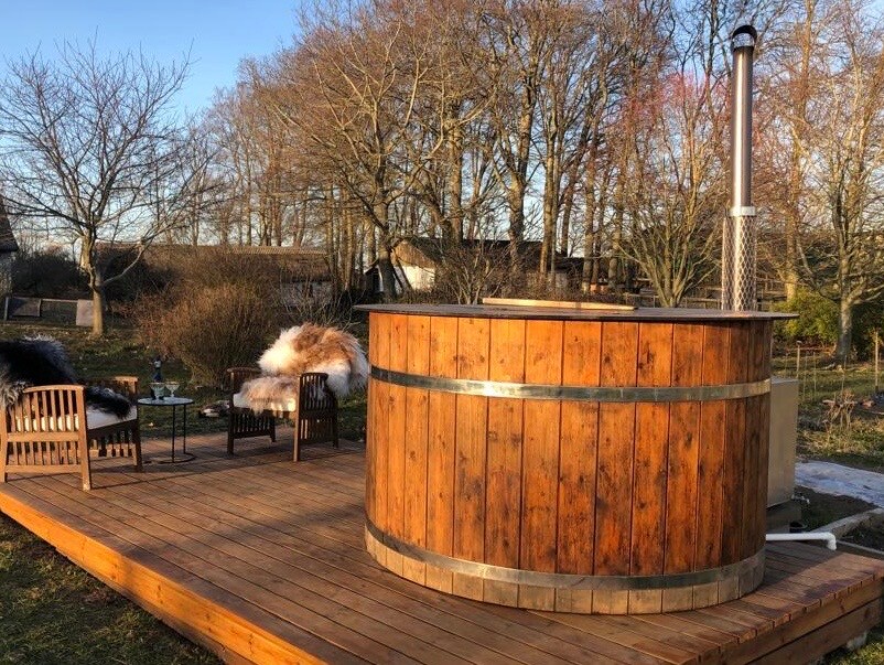 An outdoor wooden hot tub is nestled on a deck surrounded by trees. Two comfortable chairs are placed nearby, with cozy throws draped over them. Natural scenery extends in the background, creating a peaceful setting for relaxation.