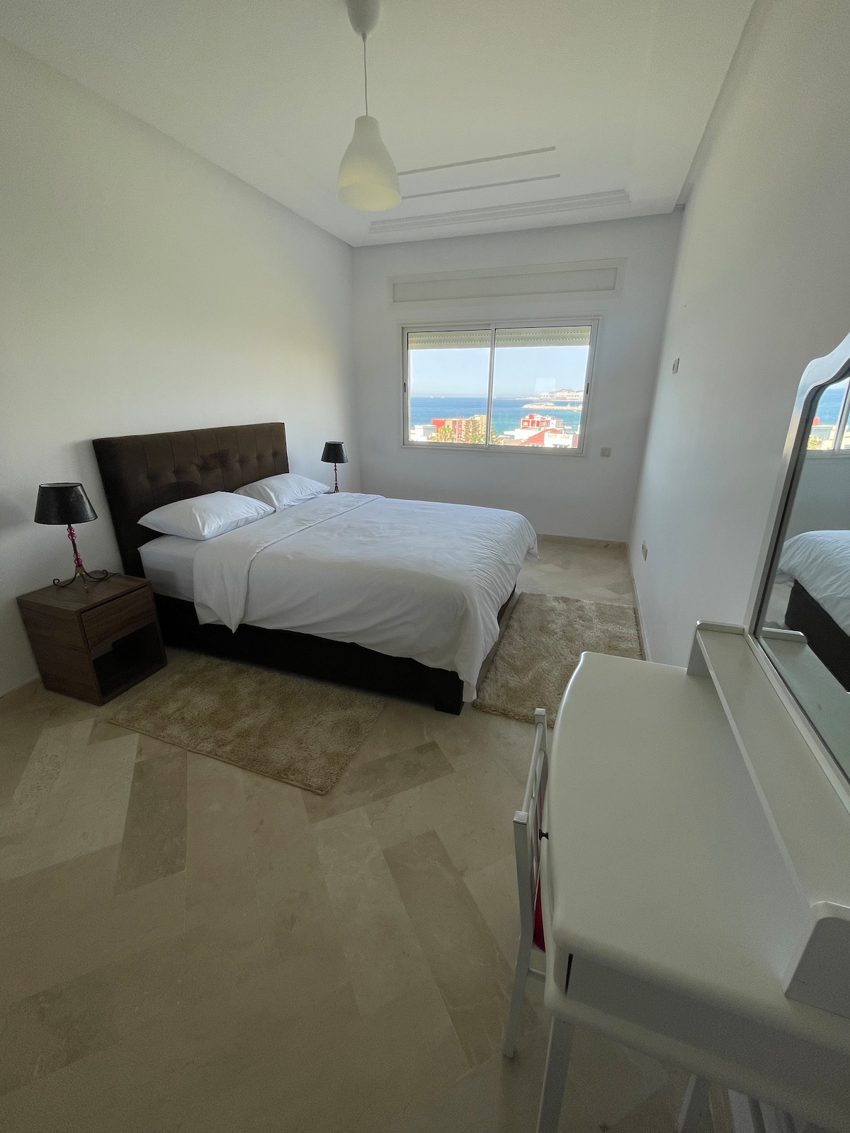 A serene bedroom contains a spacious bed with a dark upholstered headboard, complemented by bedside tables with lamps. A window offers a view of the sea, while a light-colored rug lies beneath. A vanity table with a mirror is positioned by the wall.