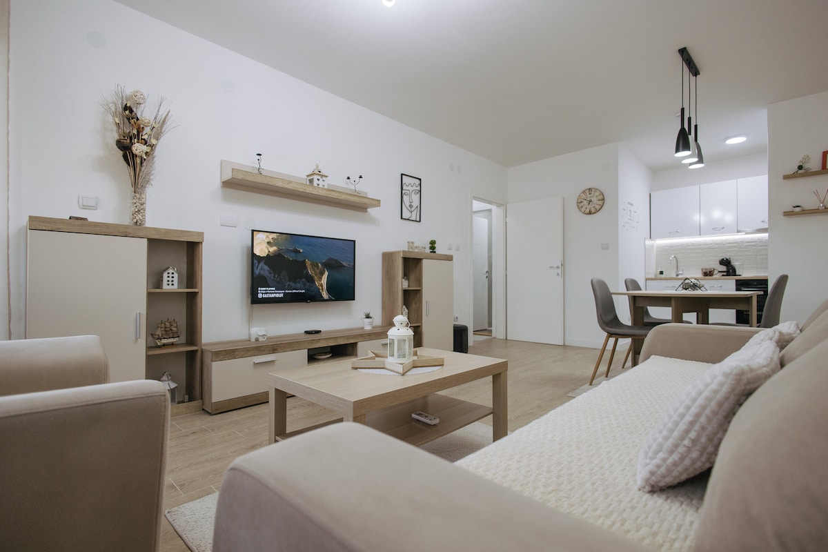 A modern living area features a light-colored sofa and a coffee table, complemented by a wooden entertainment unit housing a television. A dining area with a table and chairs is visible in the background. Soft lighting creates a calm ambiance throughout the space.