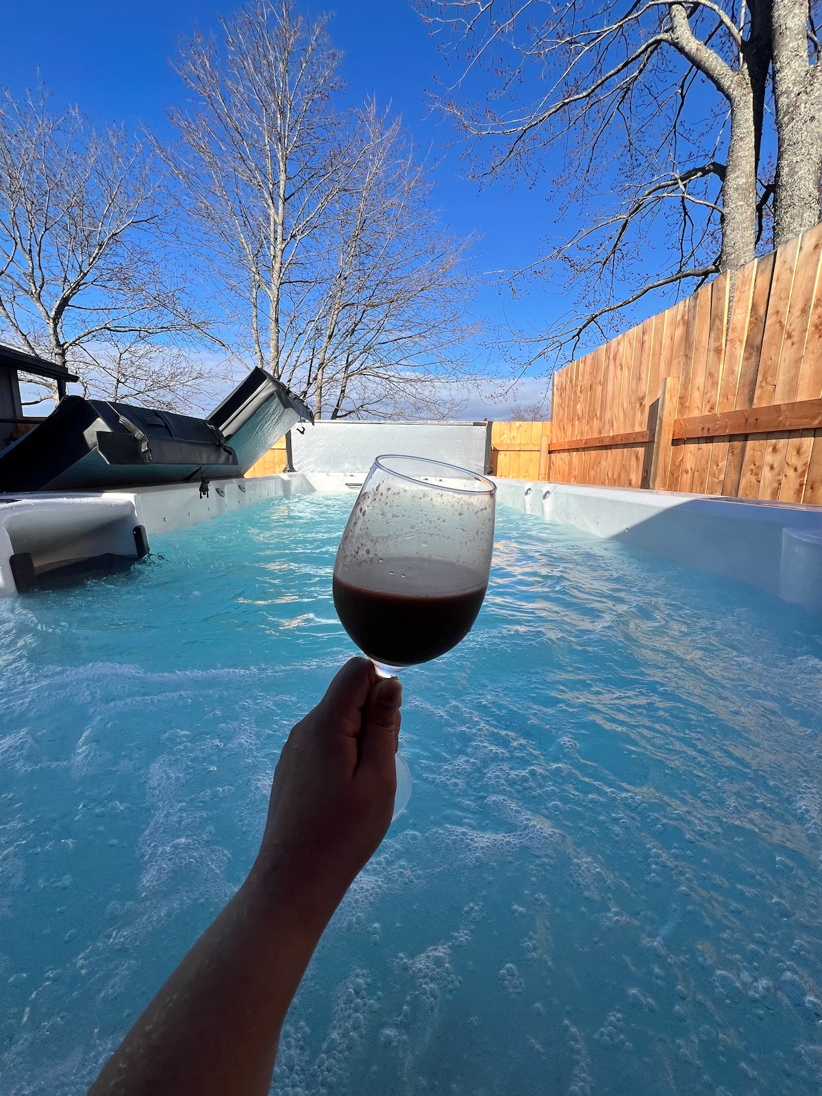 A relaxing hot tub is set against a backdrop of clear blue skies and bare trees. The water's surface is calm, reflecting the surrounding wood fencing. A hand is seen holding a glass, indicating a peaceful moment of enjoyment.
