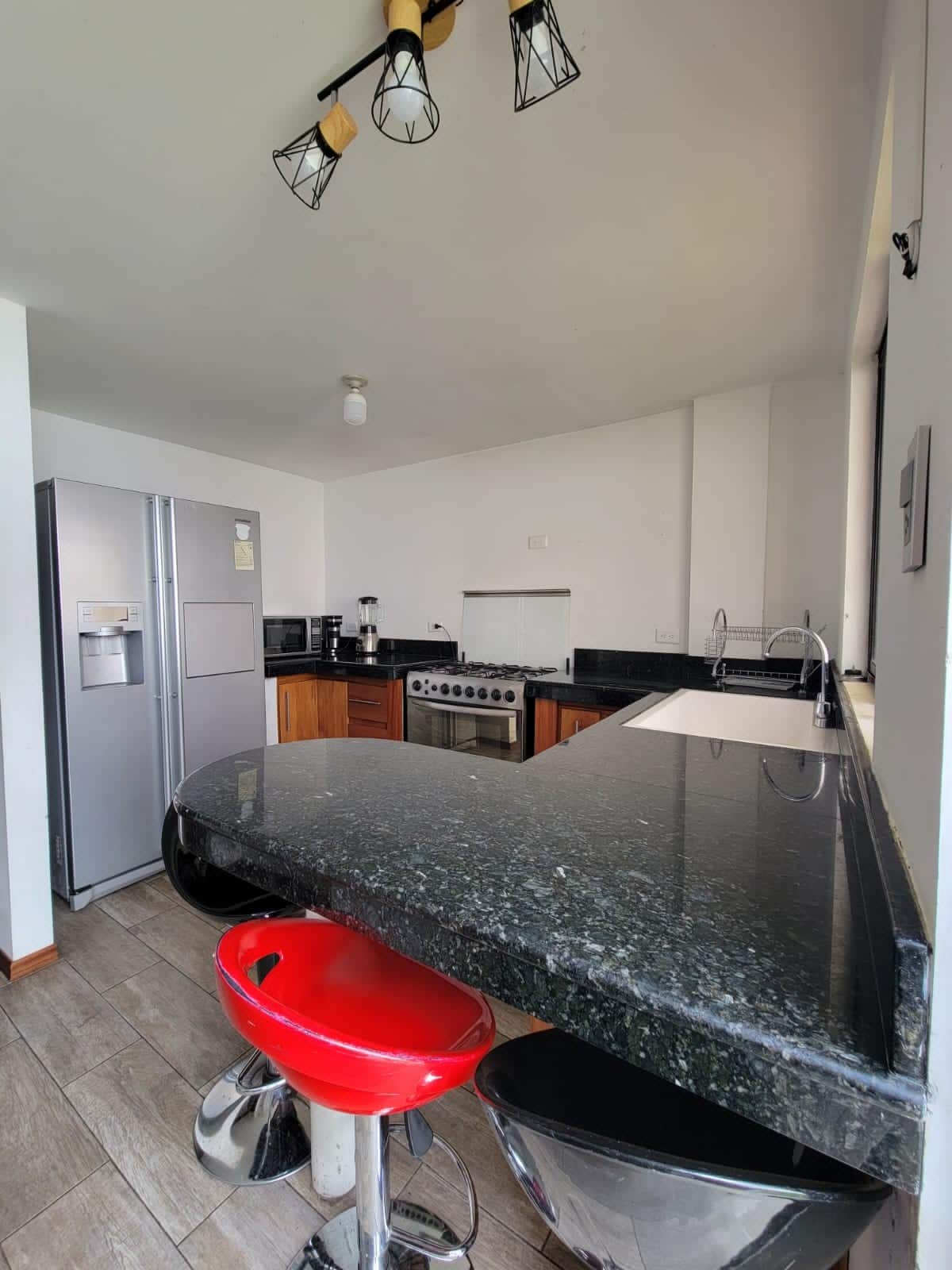 The kitchen features sleek granite countertops with a dark finish and wooden cabinetry. A modern stove and a large refrigerator are visible, along with a coffee maker and microwave. A curved bar area with red stools enhances the space, illuminated by stylish overhead lights.