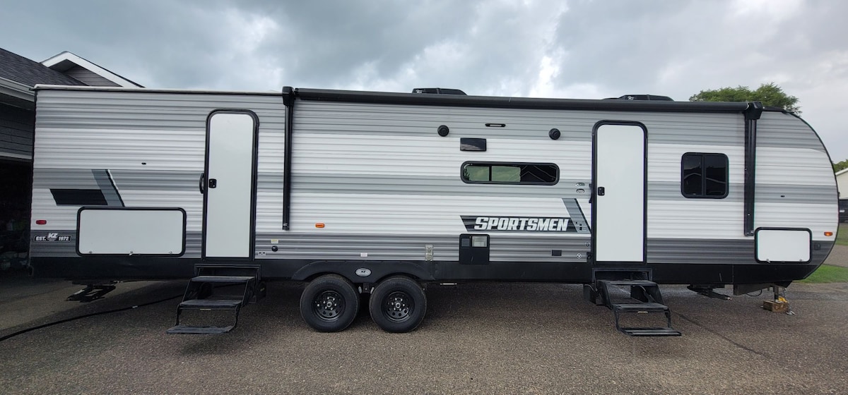 A spacious camper is shown, featuring a sleek silver exterior with black accents. Two entrance doors are visible, providing convenient access. Large windows are strategically placed, allowing natural light to enter. The dual-axle design ensures stability and easy maneuverability when traveling.