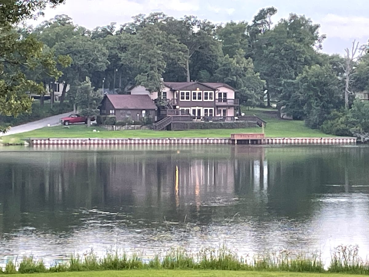 The image shows a two-story duplex set against a lush green landscape, situated by a calm lake. The building features a large deck with multiple windows facing the water, reflecting the surrounding trees. A wooden dock extends into the lake, enhancing the serene setting.