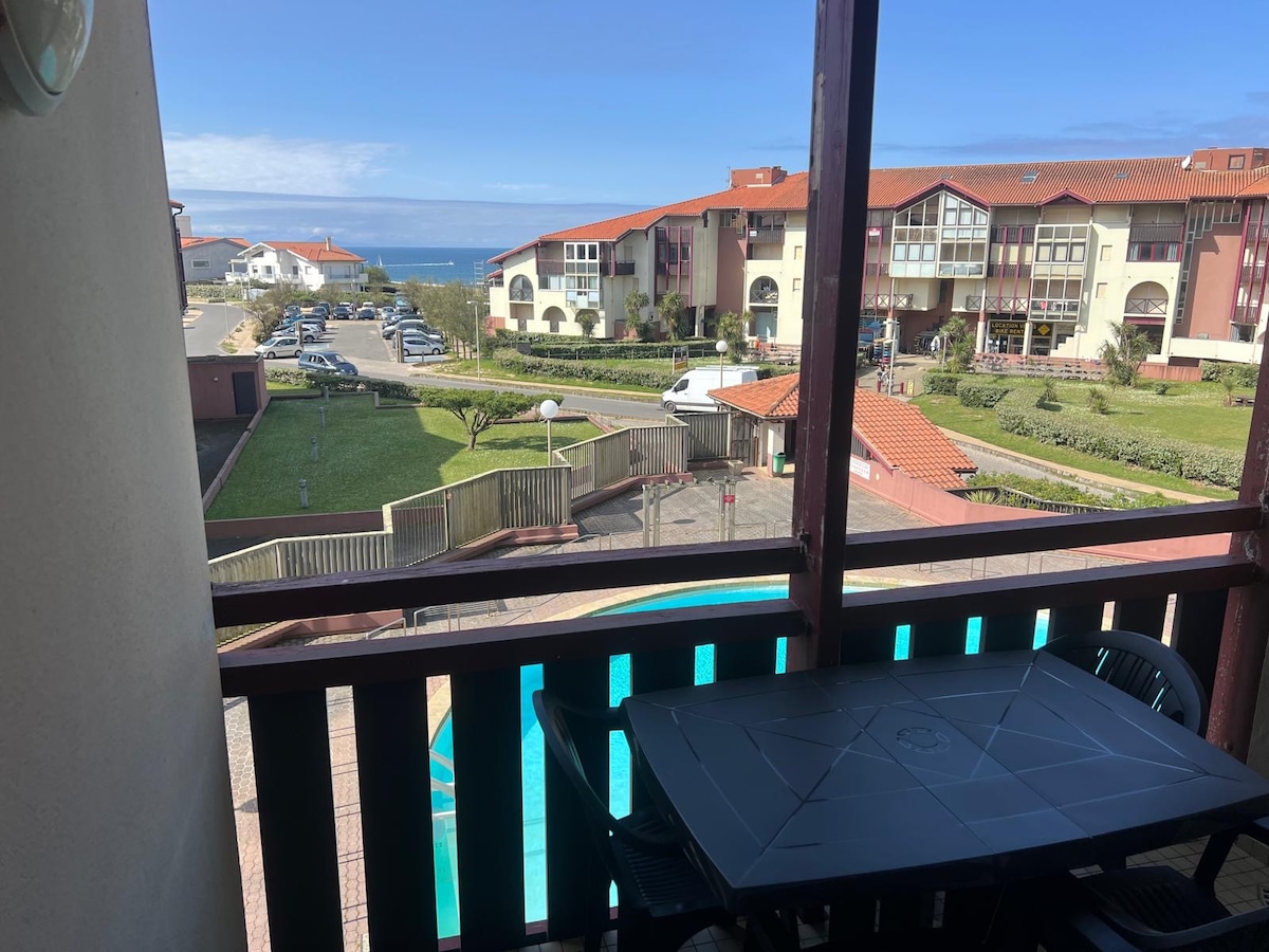 A terrace view overlooks the pool and residential grounds, with the ocean visible in the background. A table with four chairs is positioned on the terrace, inviting outdoor dining. The scenic surroundings and well-maintained landscaping can be appreciated from this spot.
