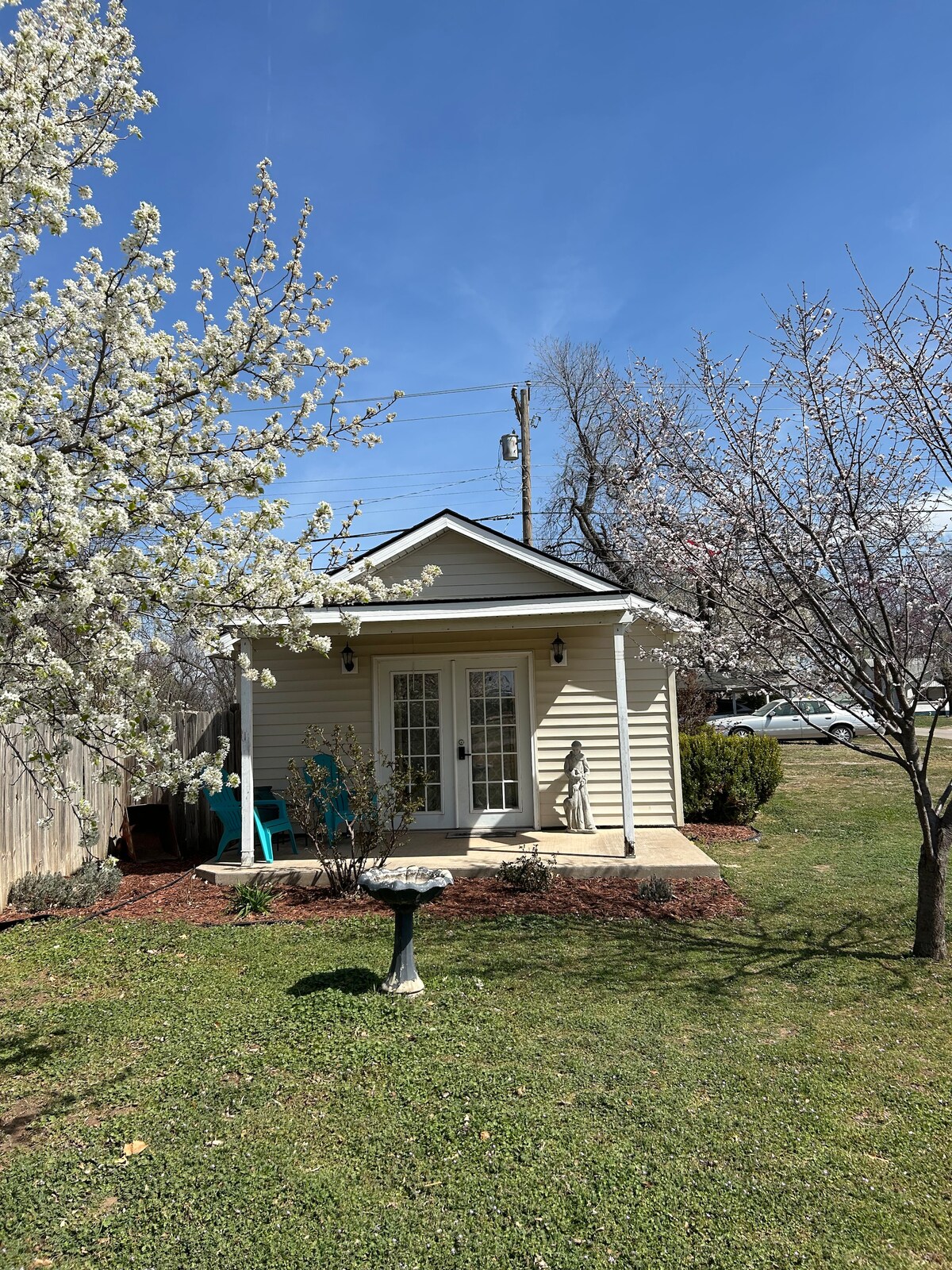 A charming exterior of a small one-room suite is highlighted by blooming trees and freshly mowed grass. French doors provide access to the entryway, while a birdbath and two turquoise chairs add to the inviting outdoor space. The sky is clear and blue.