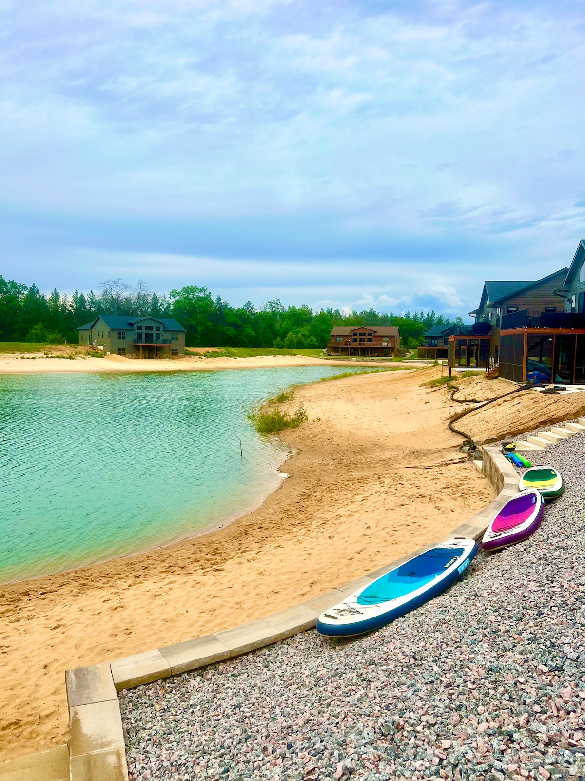 A serene lakeside view features a sandy beach with gentle waves lapping at the shore. Three colorful stand-up paddle boards are lined up along the textured stone path, with lush green trees framing the tranquil setting, creating an inviting space for relaxation and outdoor activities.