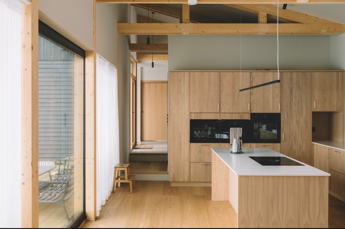 A spacious kitchen area is presented with light wooden cabinetry and a central island featuring a sleek countertop. Large glass doors allow natural light to flood the space, leading to an outdoor seating area. The minimalist design emphasizes clean lines and a serene environment.
