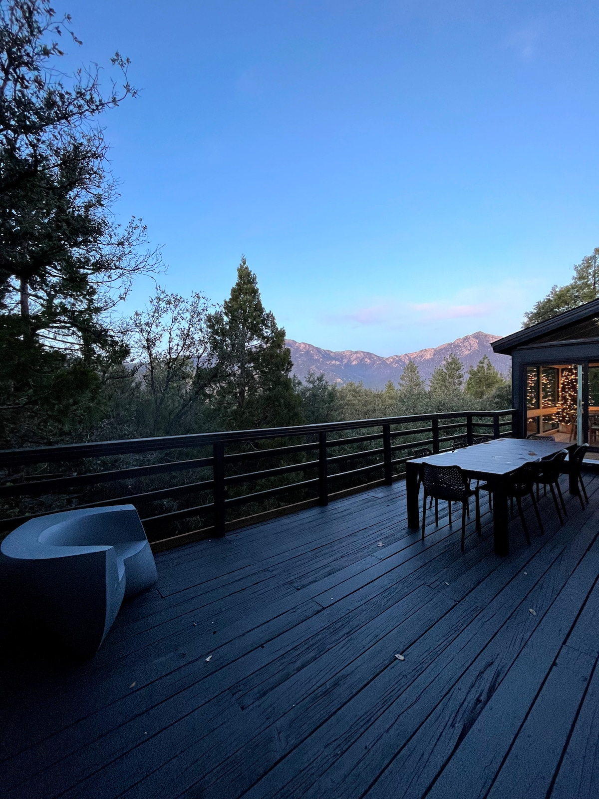An expansive outdoor deck features a large dining table surrounded by chairs, offering views of the San Jacinto mountains. A modern chair is positioned nearby, and the deck is adorned with a wooden railing, seamlessly blending with the natural surroundings.
