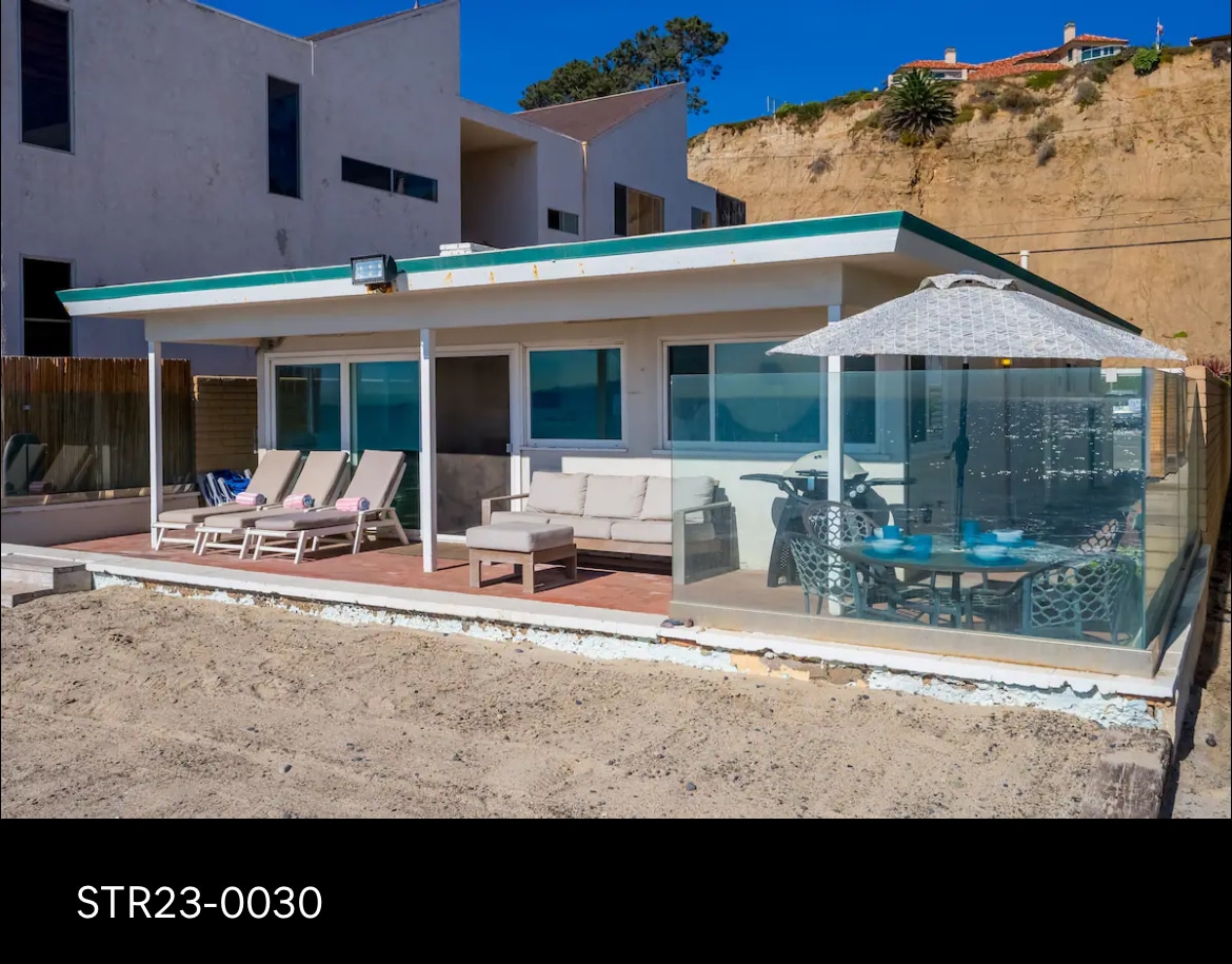 A beachfront patio area is visible, featuring a set of loungers and a seating arrangement. A dining table with chairs is located under a shaded umbrella. Glass panels provide an unobstructed view of the beach, enhancing the outdoor experience.