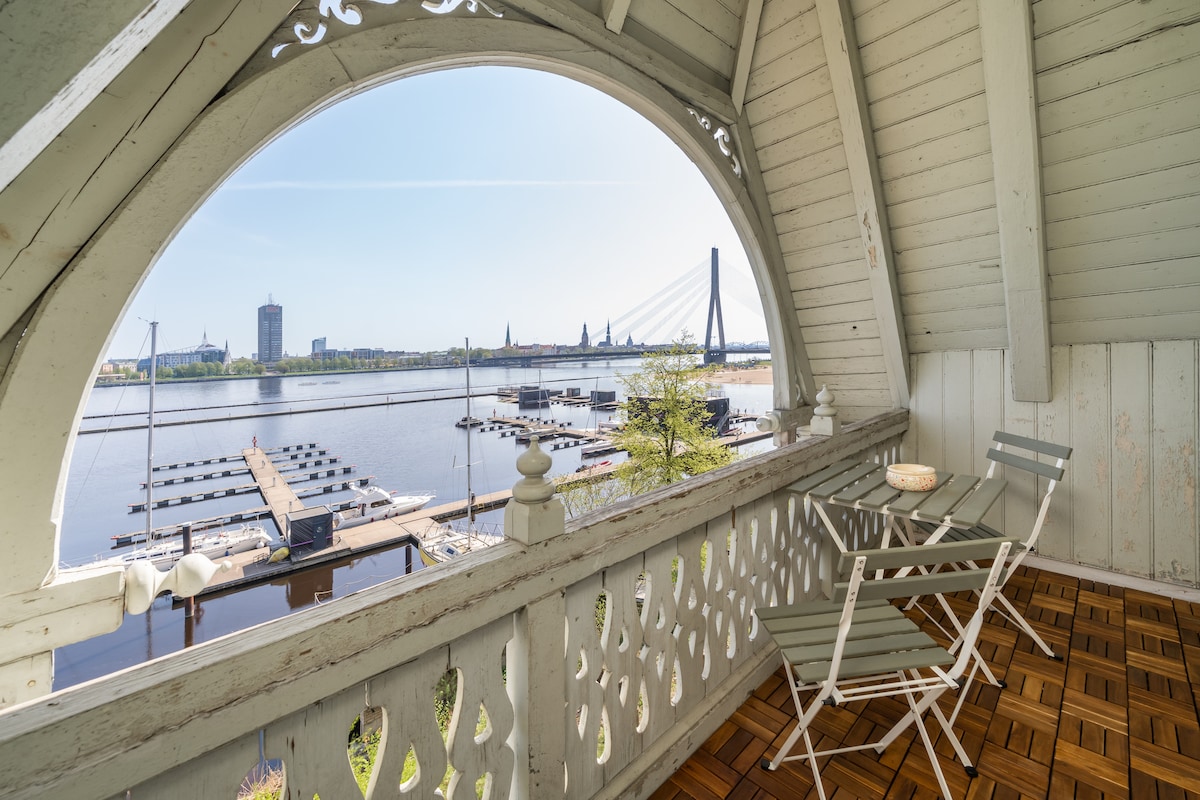 A charming balcony features wooden flooring and detailed railings, offering a scenic view of the river and city skyline. Two white metal chairs are positioned for relaxation, overlooking the marina with boats and the distinctive bridge in the background under a clear blue sky.
