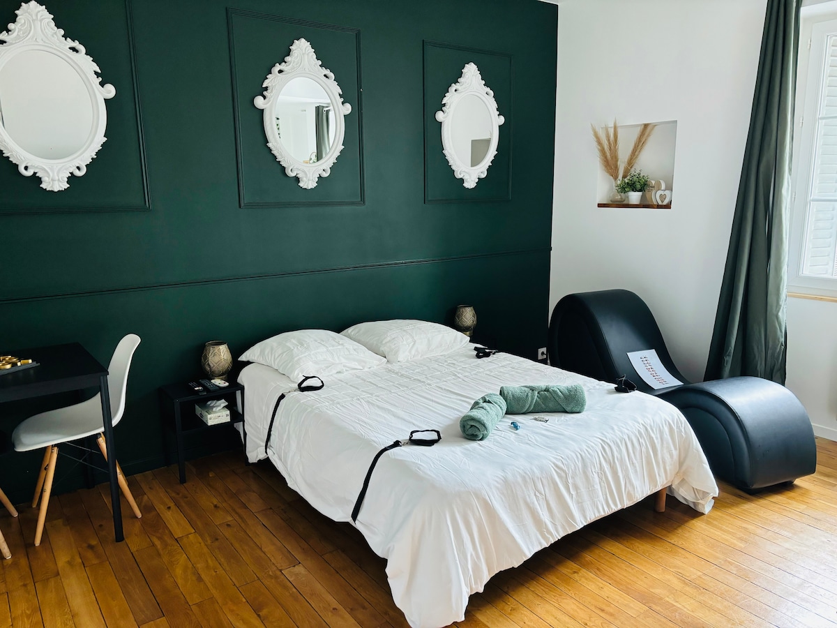 A contemporary sleeping area features a spacious bed dressed in white linens. Three ornate mirrors are mounted on a dark green wall. A black chaise lounge is positioned beside the bed, and a small table with a chair is also visible in the room.