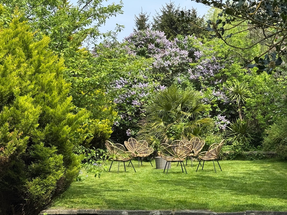 A serene garden setting is presented, featuring a green lawn surrounded by lush trees and flowering shrubs. Six wicker chairs are arranged in a circular pattern, creating an inviting space for relaxation and conversation.