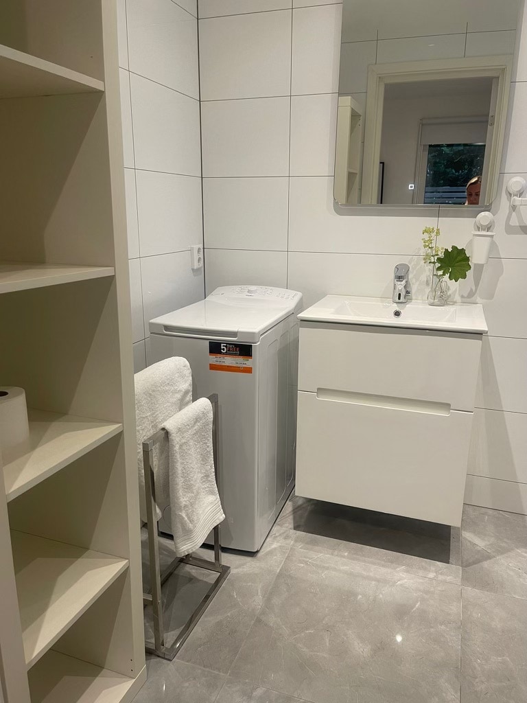 A modern bathroom is depicted, featuring a compact washing machine next to a sleek vanity. Soft towels are neatly arranged on a rack, while a small plant adds a touch of greenery. Bright lighting reflects off the tiled walls and floor, enhancing the clean design.