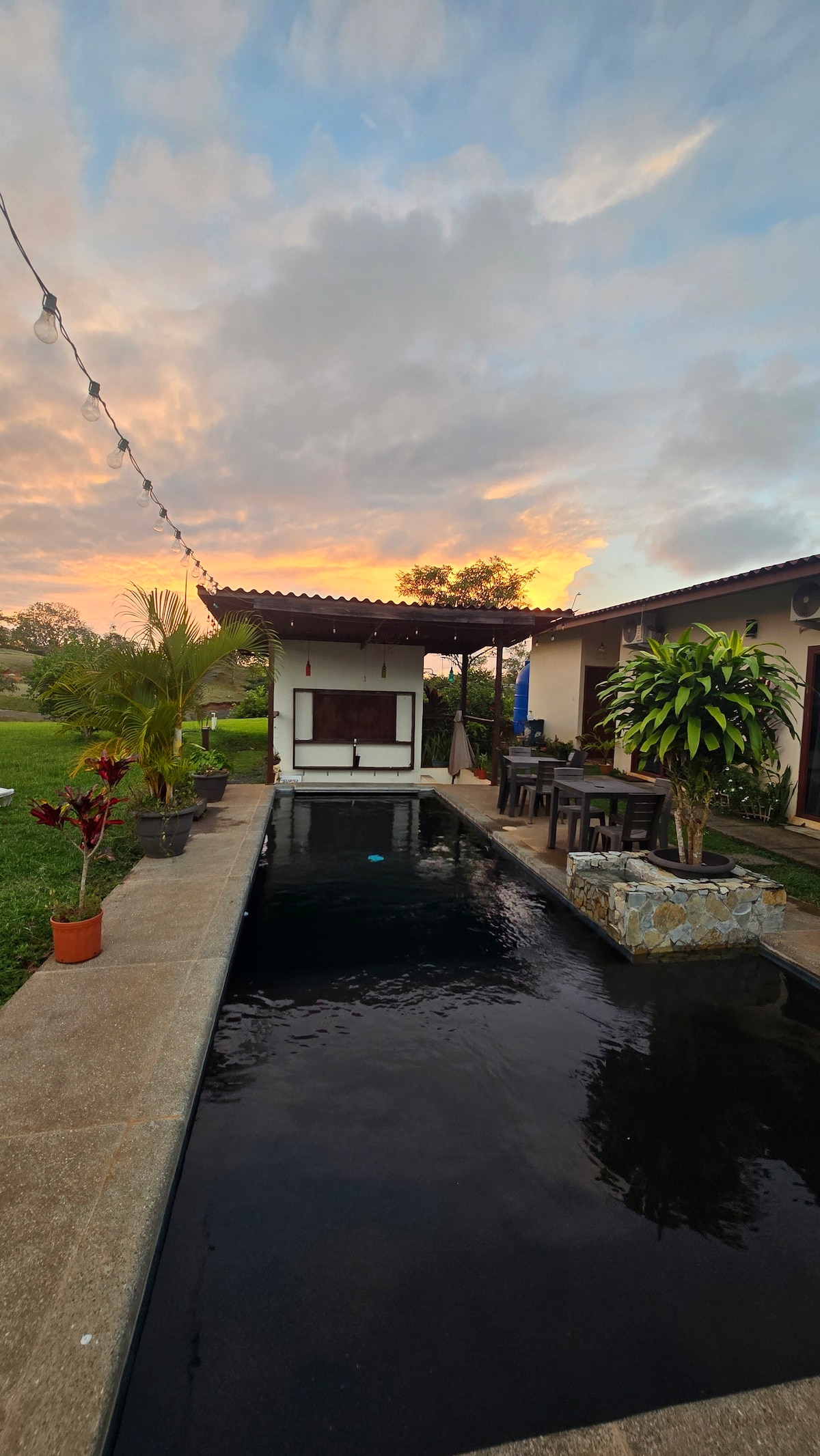 A tranquil courtyard is depicted with a dark pool reflecting the evening sky. Surrounding greenery adds a natural touch. A covered seating area is present nearby, accompanied by a dining table. String lights above create a gentle ambiance as the sun sets on the horizon.