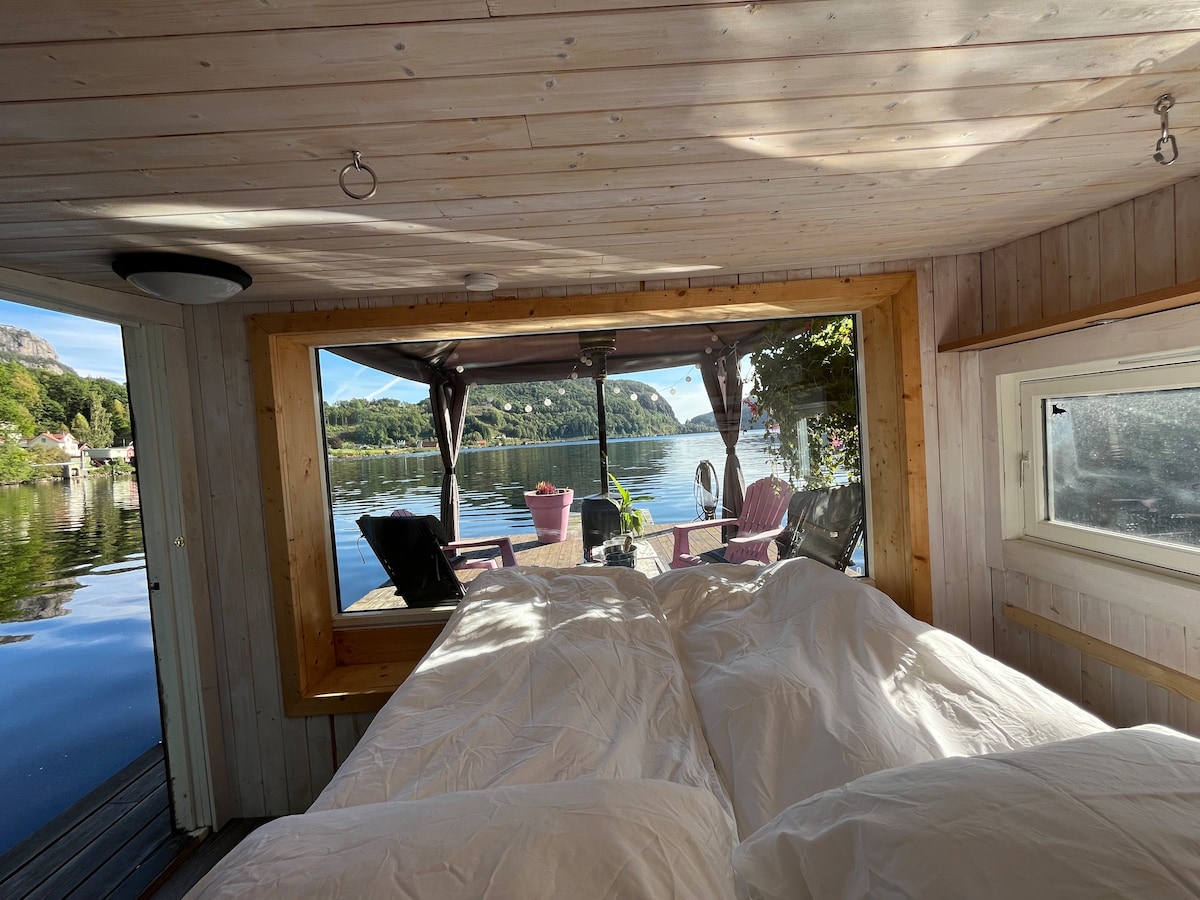 A tranquil sleeping area features a spacious bed covered with white bedding. A large window offers a scenic view of the calm water and surrounding landscape. Outdoor seating is visible nearby, enhancing the serene lakeside atmosphere.