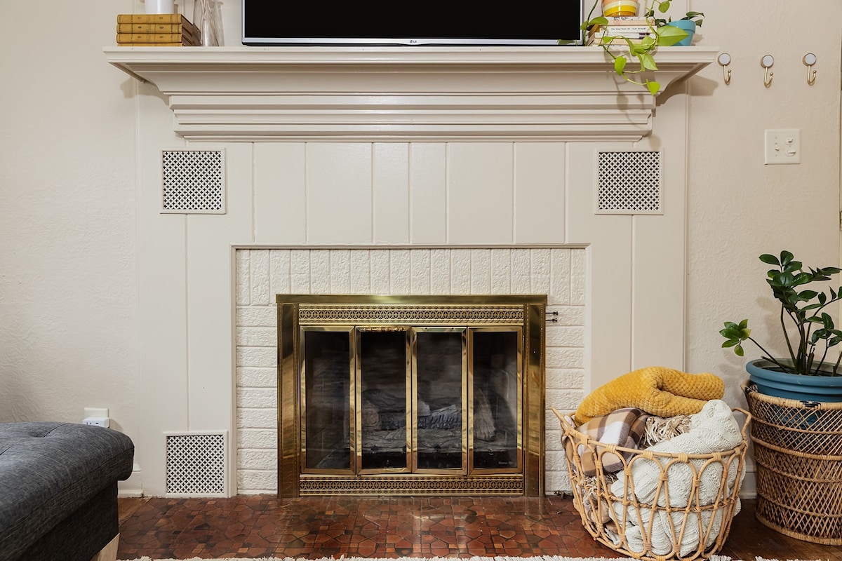 A decorative fireplace is framed by white paneling, featuring a brass trim. The firebox is enclosed with glass doors, while a woven basket filled with colorful blankets is positioned nearby. Greenery is visible in a decorative pot on the side.