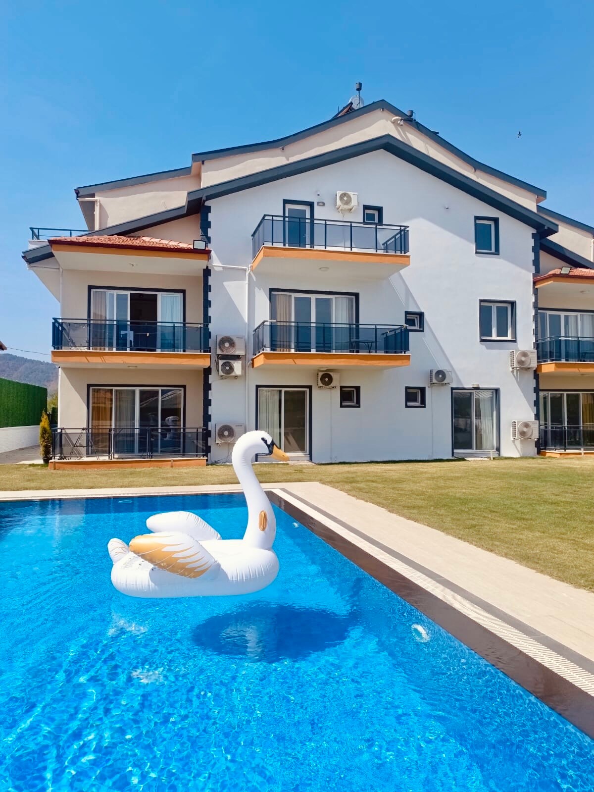 A modern building with three levels is depicted, featuring multiple balconies and large glass doors. A swan-shaped inflatable floats in a crystal-clear pool, surrounded by a well-maintained lawn. Air conditioning units are visible, ensuring comfort for guests during warmer months.