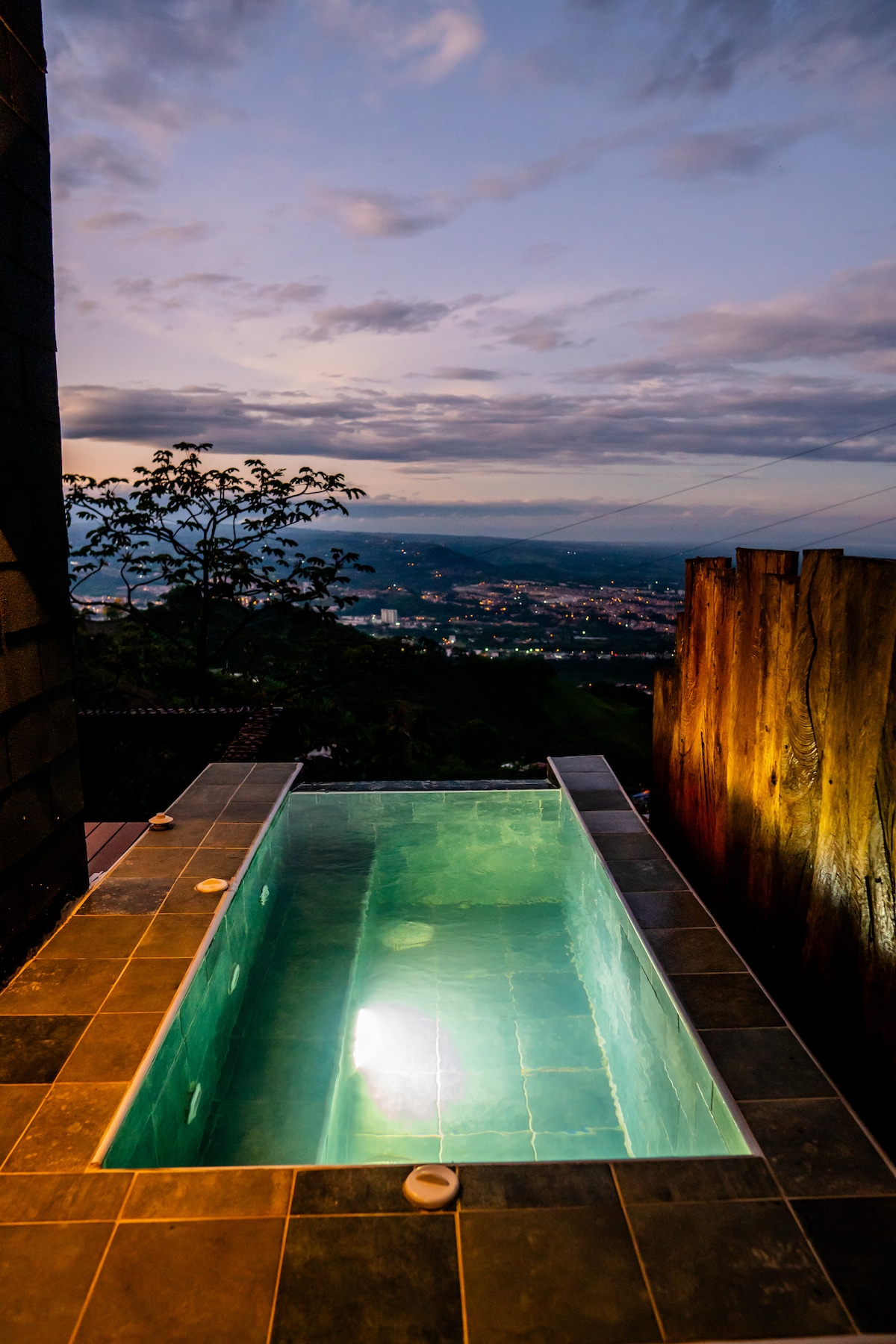 A private jacuzzi with a panoramic view is shown, set against a backdrop of a twilight sky. The water is illuminated, reflecting shades of blue and creating a serene atmosphere. Surrounding the jacuzzi is a stone patio, enhancing the outdoor experience.